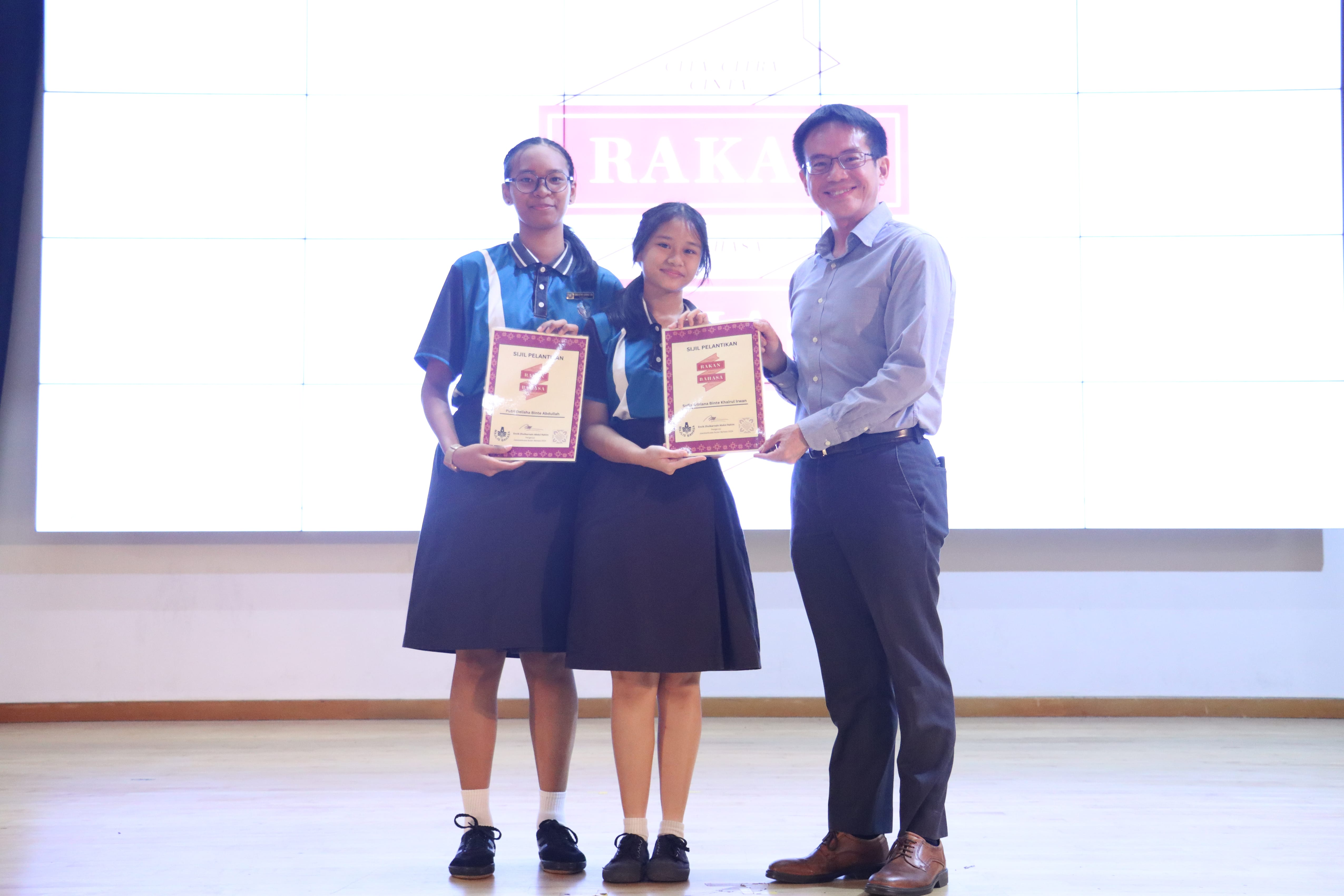 Two female students in blue uniforms hold certificates while posing with a smiling man in a gray shirt on a stage.