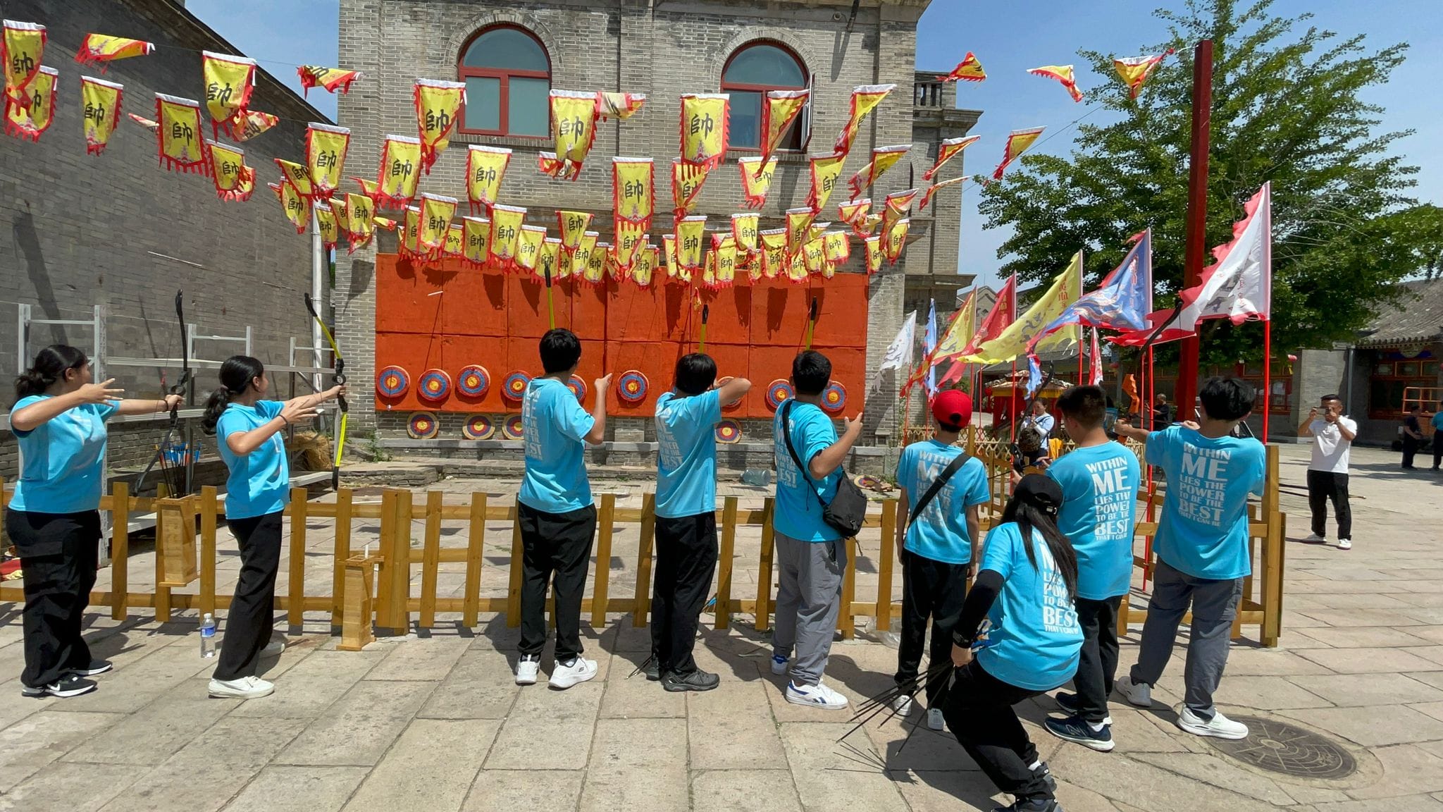 People in blue shirts practice archery at a Chinese cultural site decorated with yellow and red traditional flags.