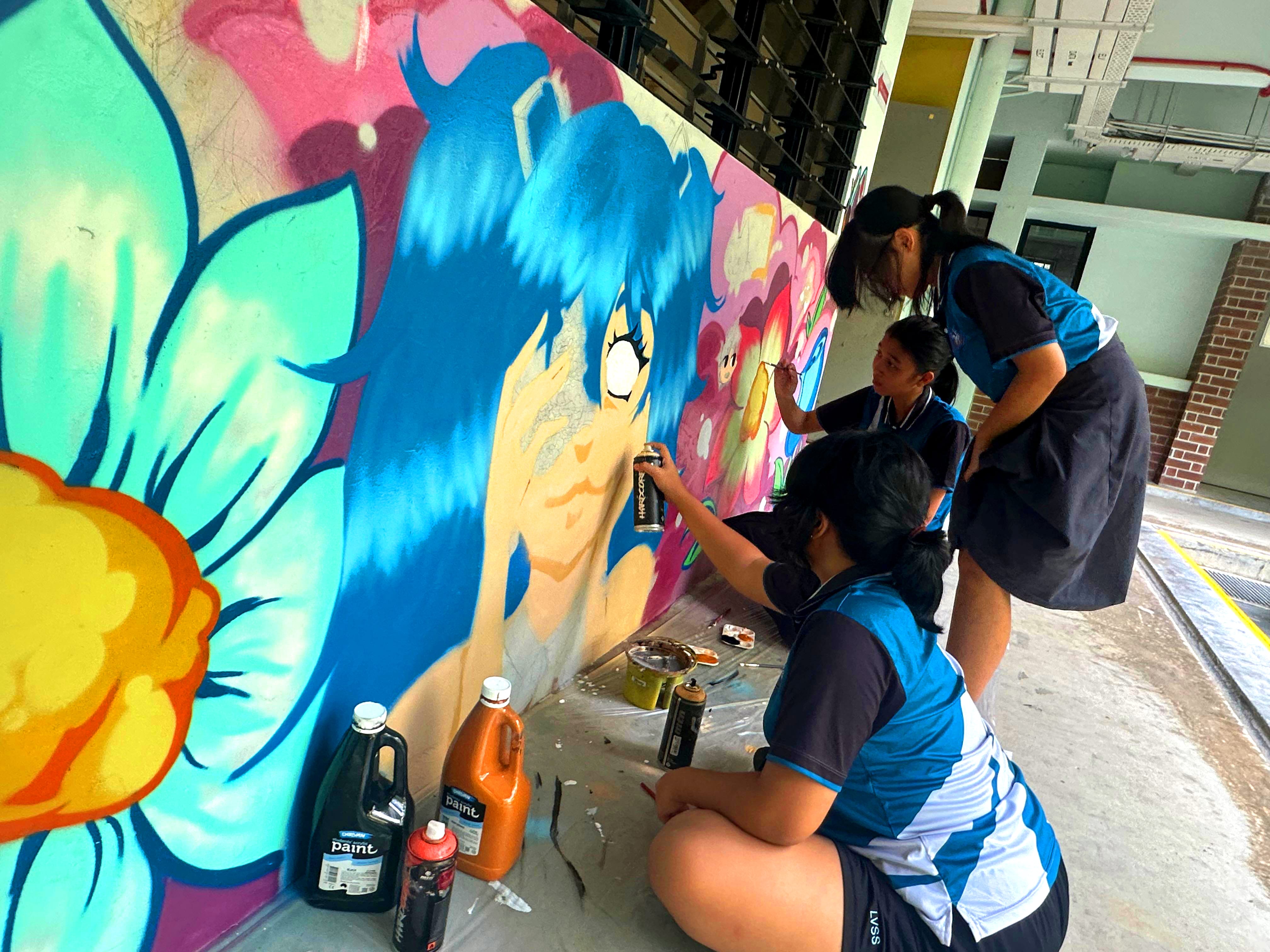 Three students paint a colorful wall mural of a blue-haired face and large flowers, with paint bottles nearby.