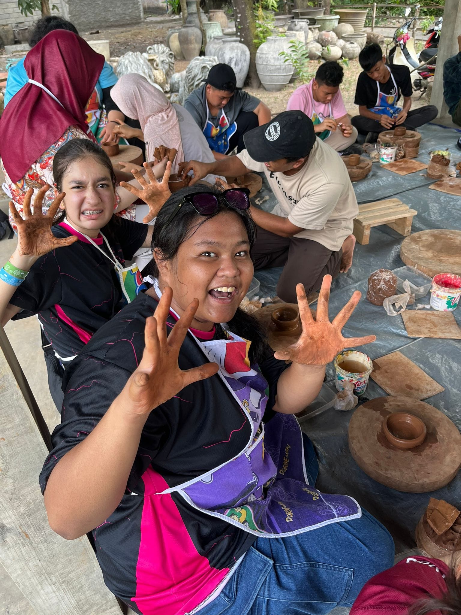 Two smiling young women show clay-covered hands during an outdoor pottery workshop with others working in the background.