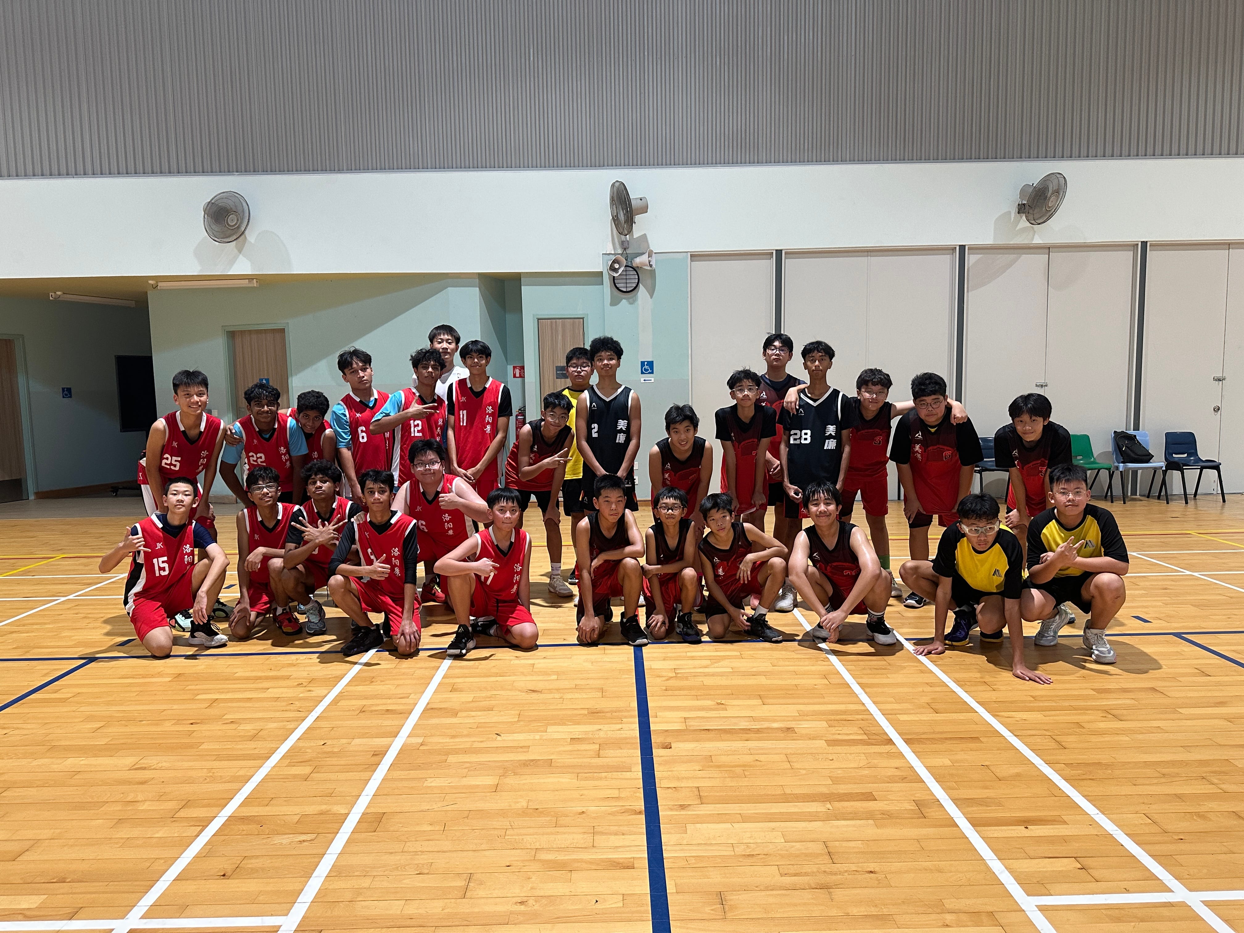 A large group of young male basketball players in red and black jerseys posing together on an indoor wooden court.