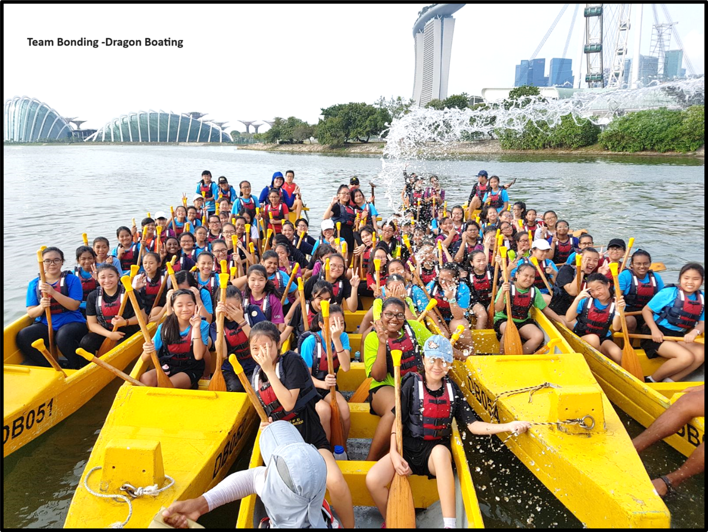 A large group of students in life jackets holding paddles sit in yellow dragon boats on a river in Singapore.