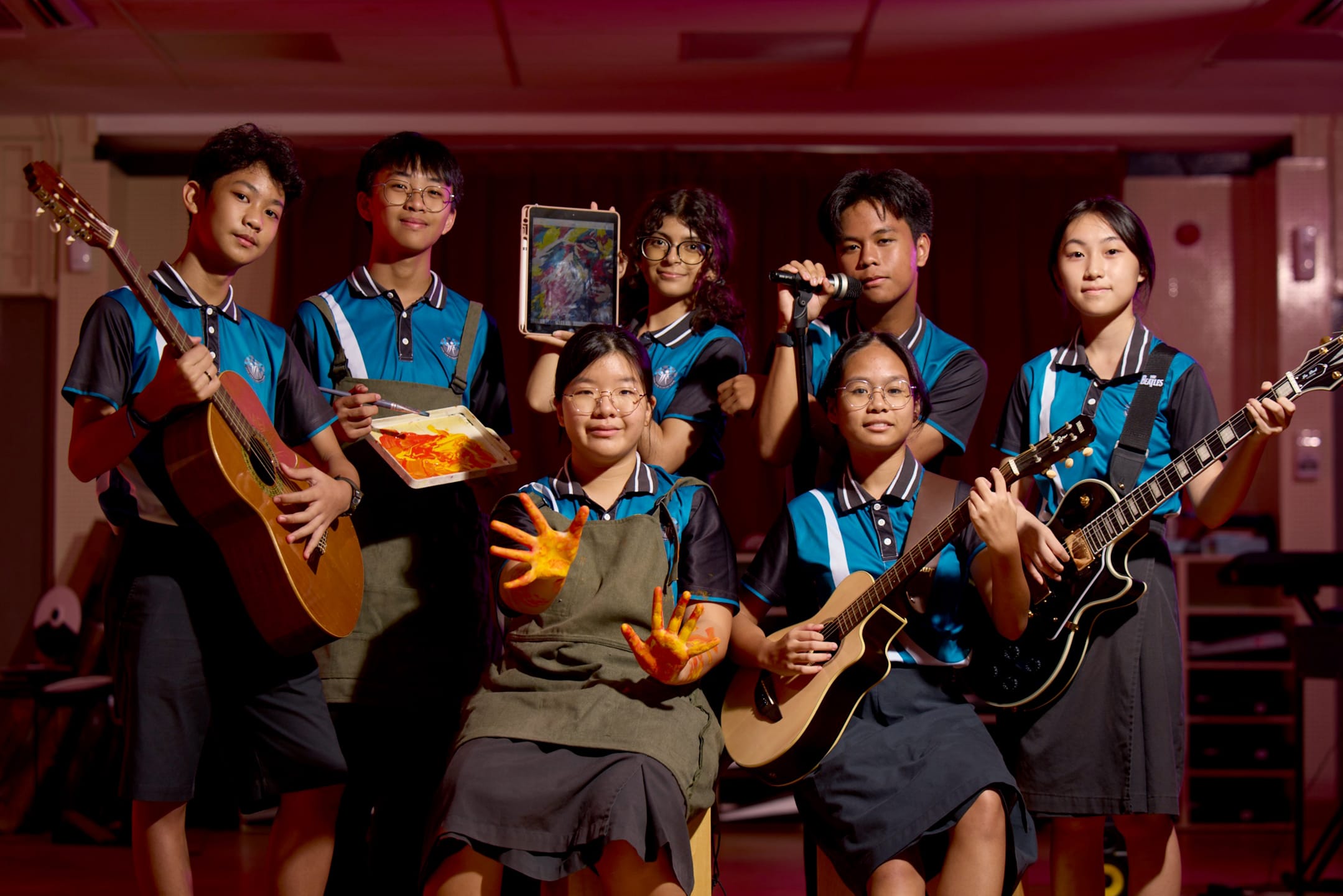 Seven students in blue uniforms pose with guitars, a microphone, artwork, a tablet, and paint-covered hands.