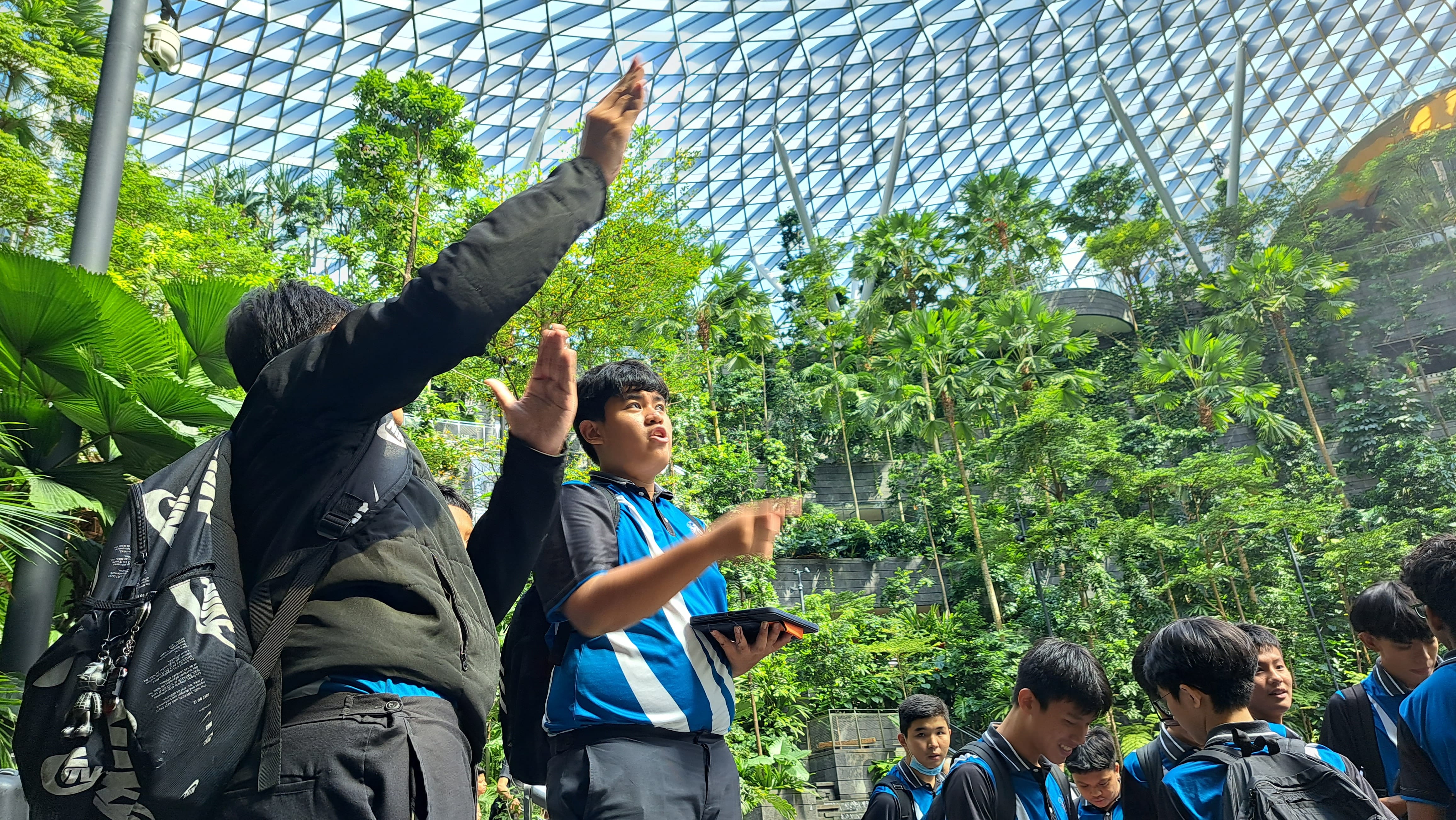 Students in blue uniforms talk and gesture under a glass dome in a lush indoor garden.