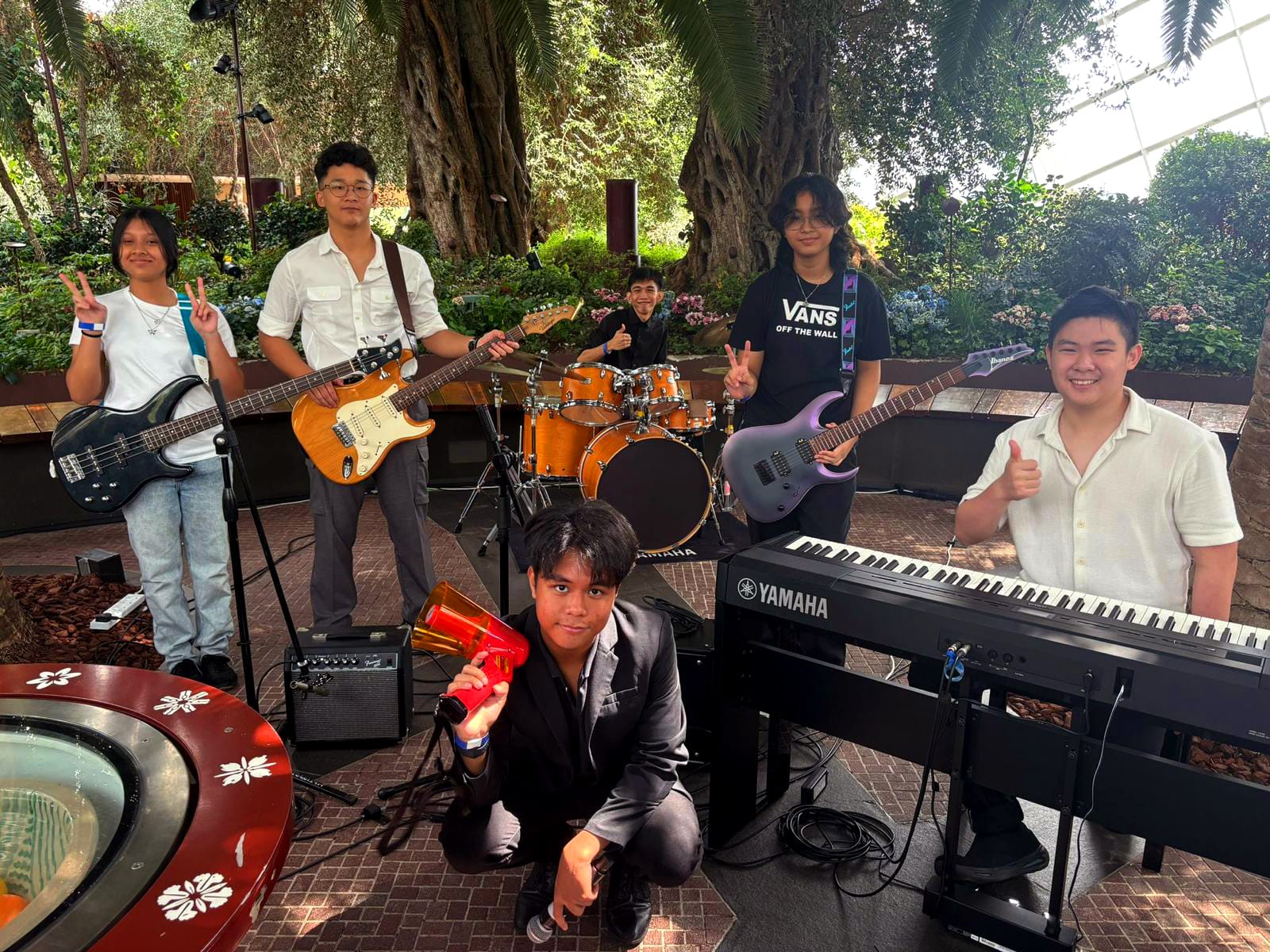 Six young musicians pose with guitars, drums, and a Yamaha keyboard in a lush indoor garden setting.