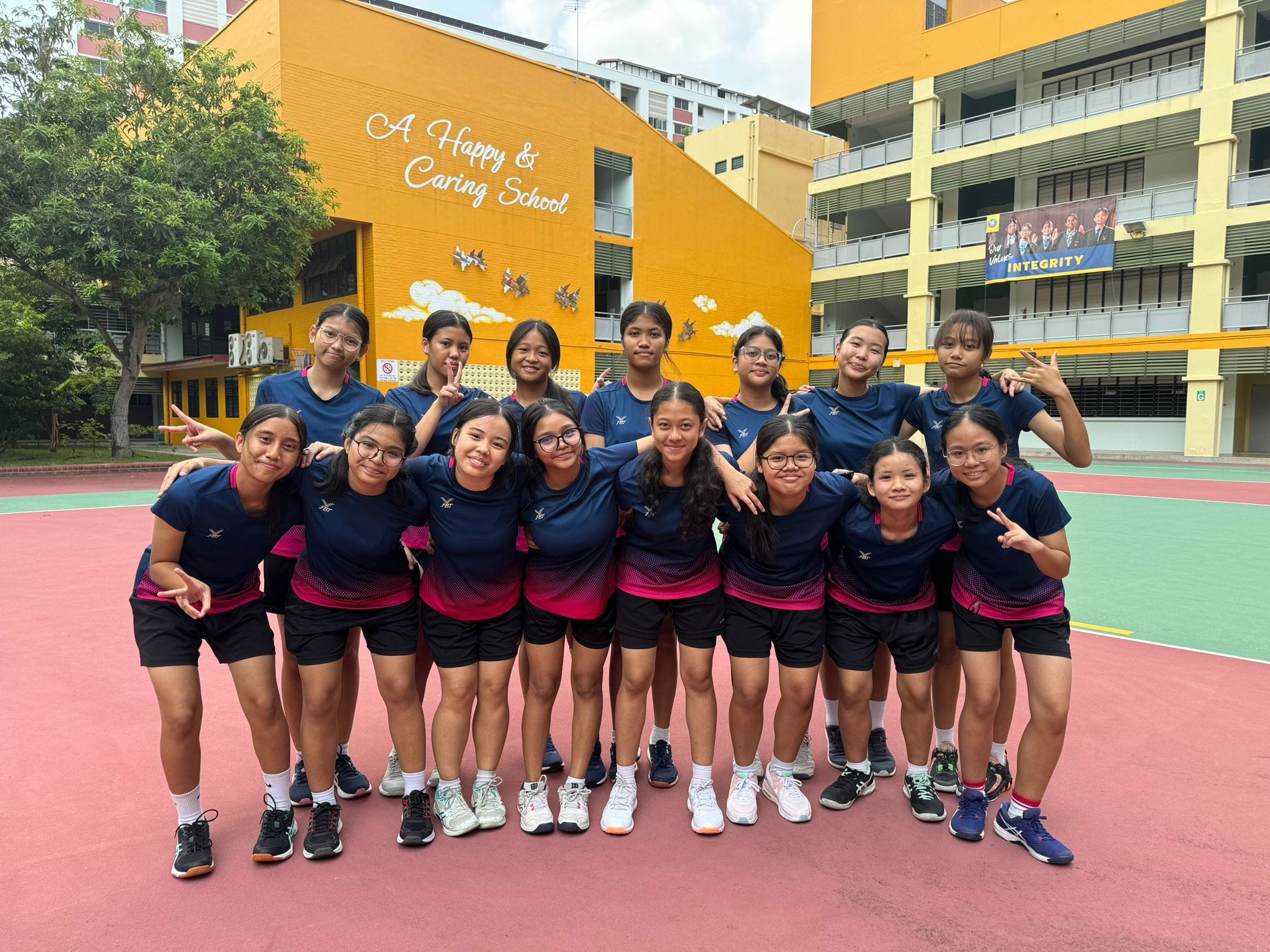 Group of teenage girls in matching navy and pink sports uniforms posing on a school court in front of a yellow building.