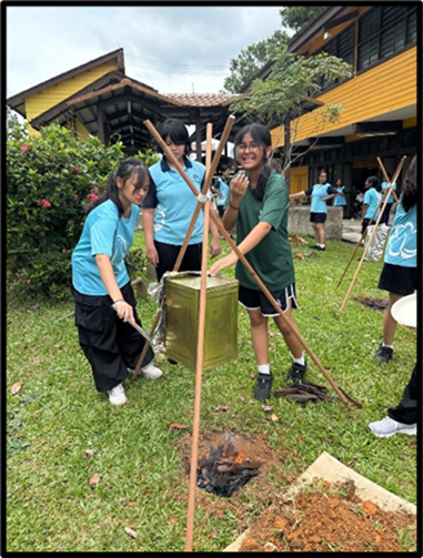 Students in blue shirts use bamboo poles to hold a container over a small fire outdoors near a yellow building.