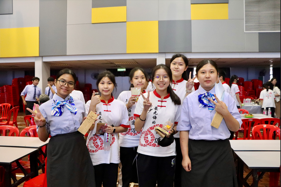 Six young female students in uniforms pose with peace signs and smiles in a hall with red chairs.