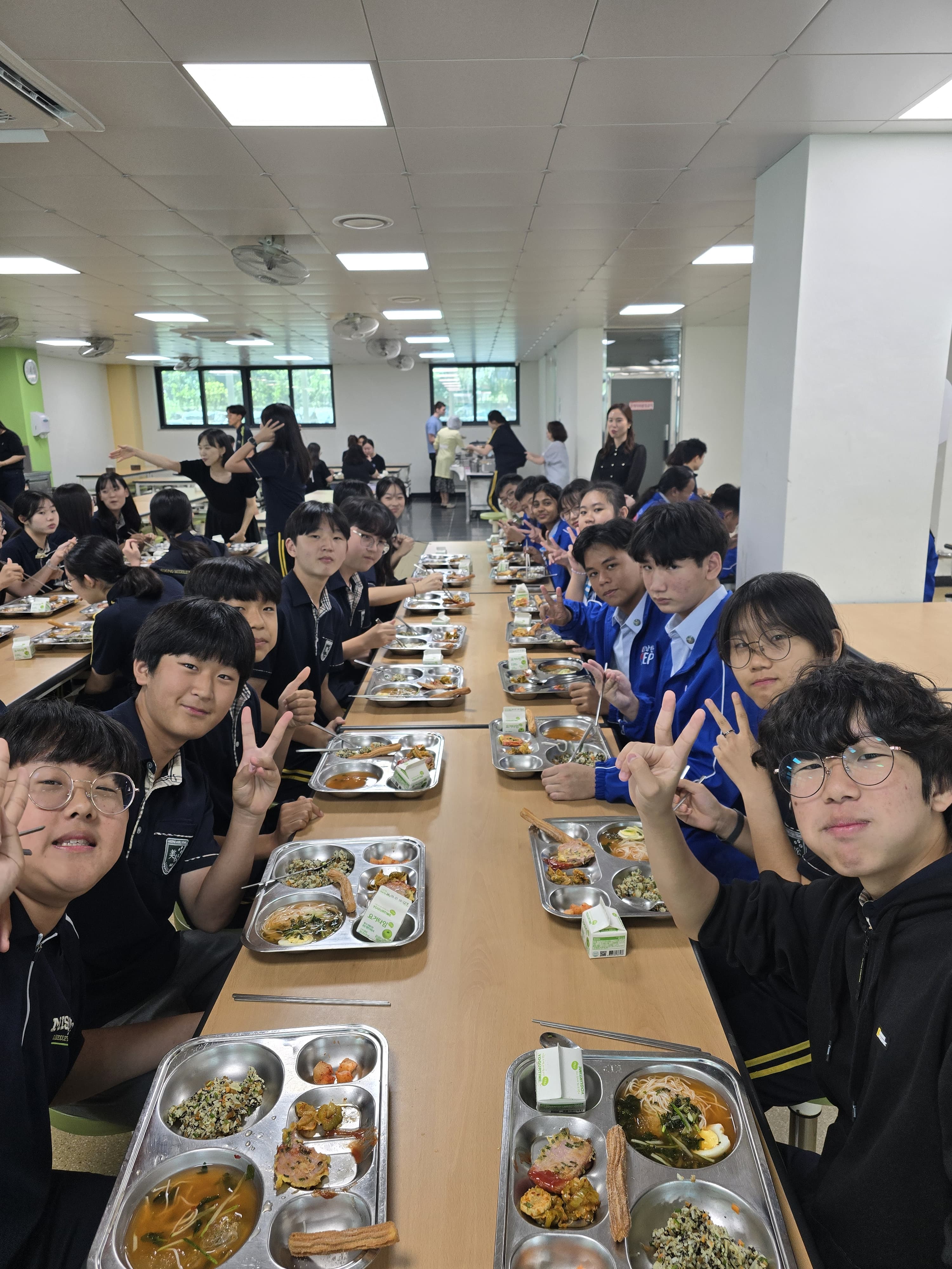 Students in uniforms sit along a long cafeteria table with metal lunch trays, many smiling and making peace signs.
