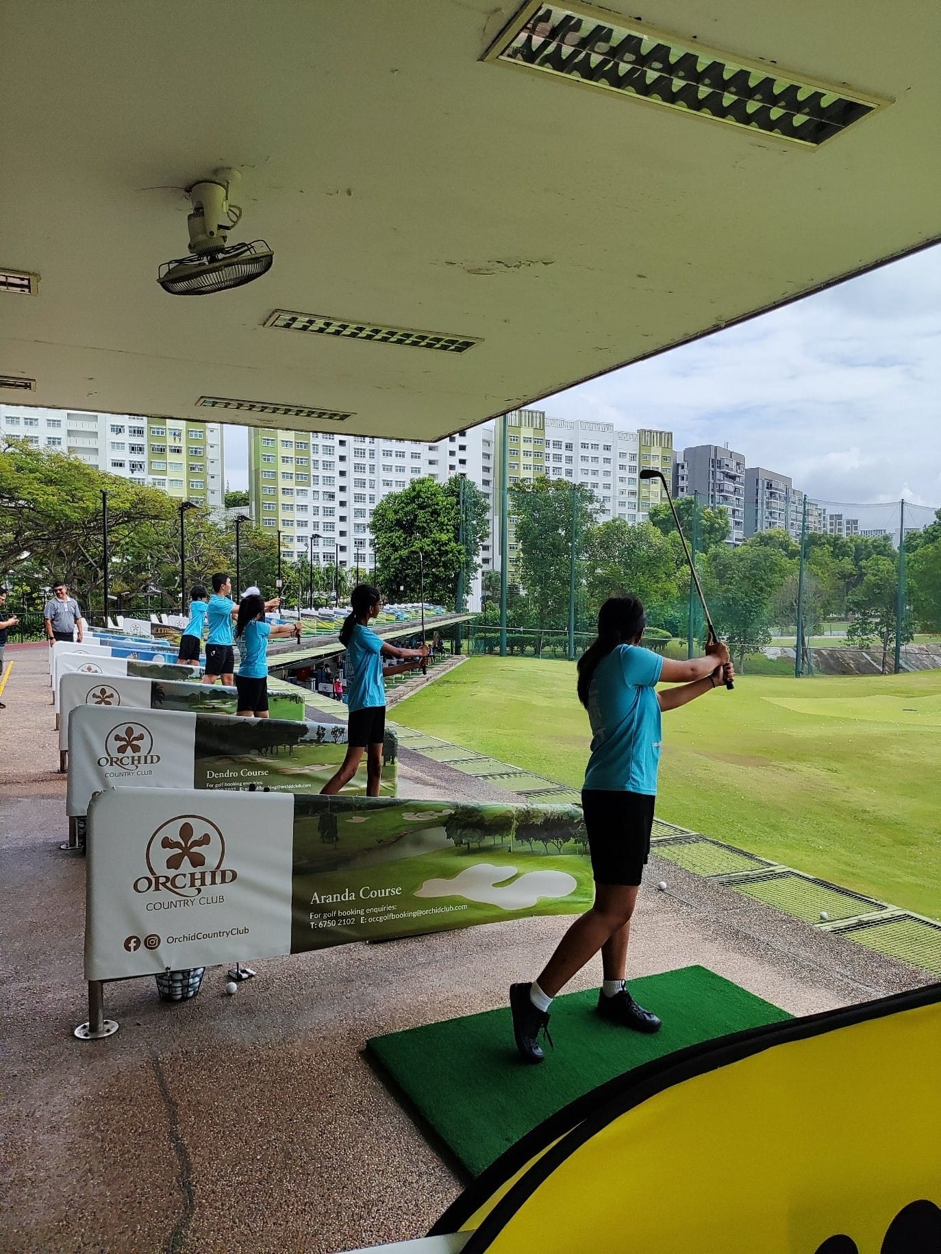 Several people in blue shirts practice golf swings at Orchid Country Club driving range with city buildings in the background.