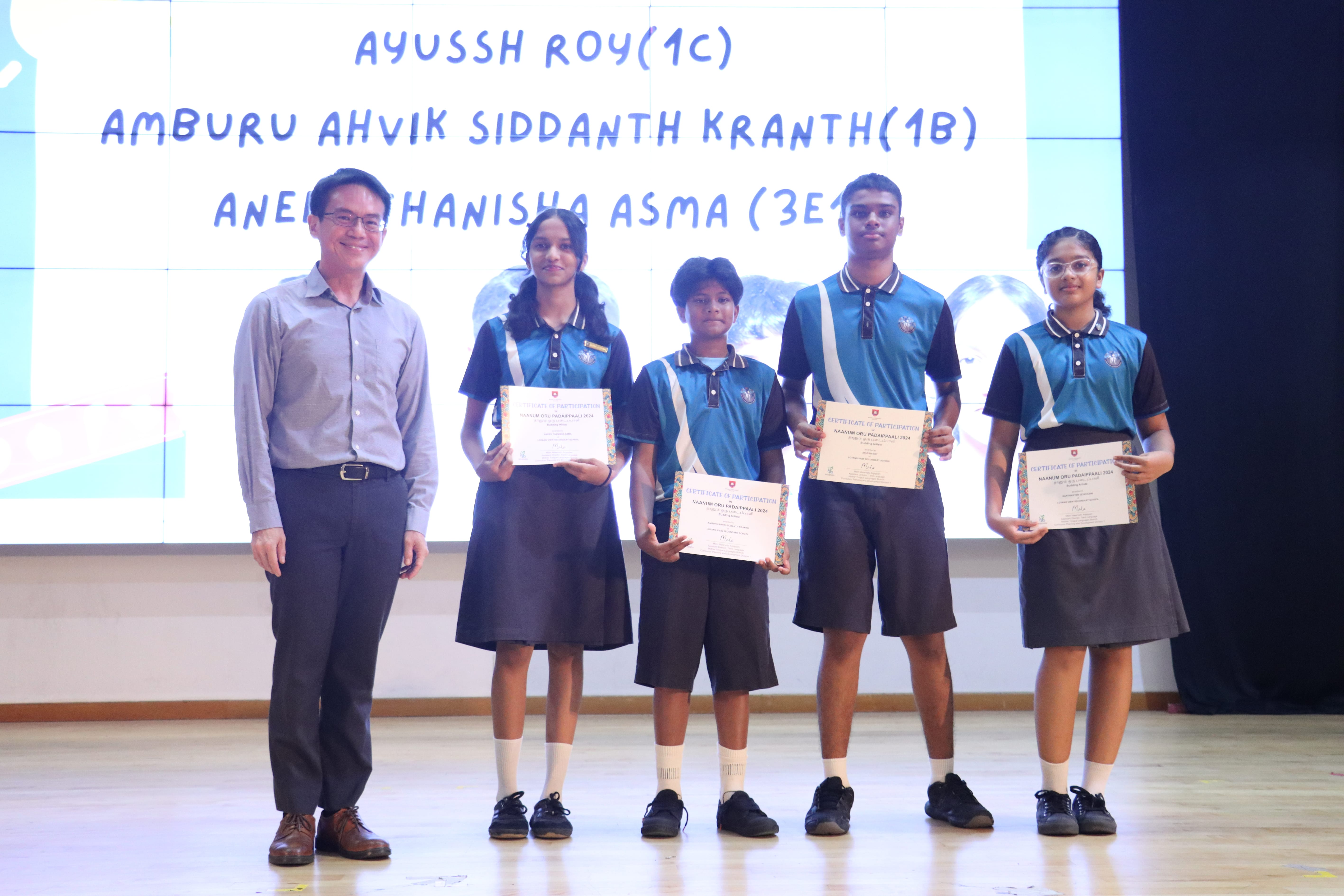 Teacher stands with four students in uniform holding certificates on stage, names displayed on screen behind.
