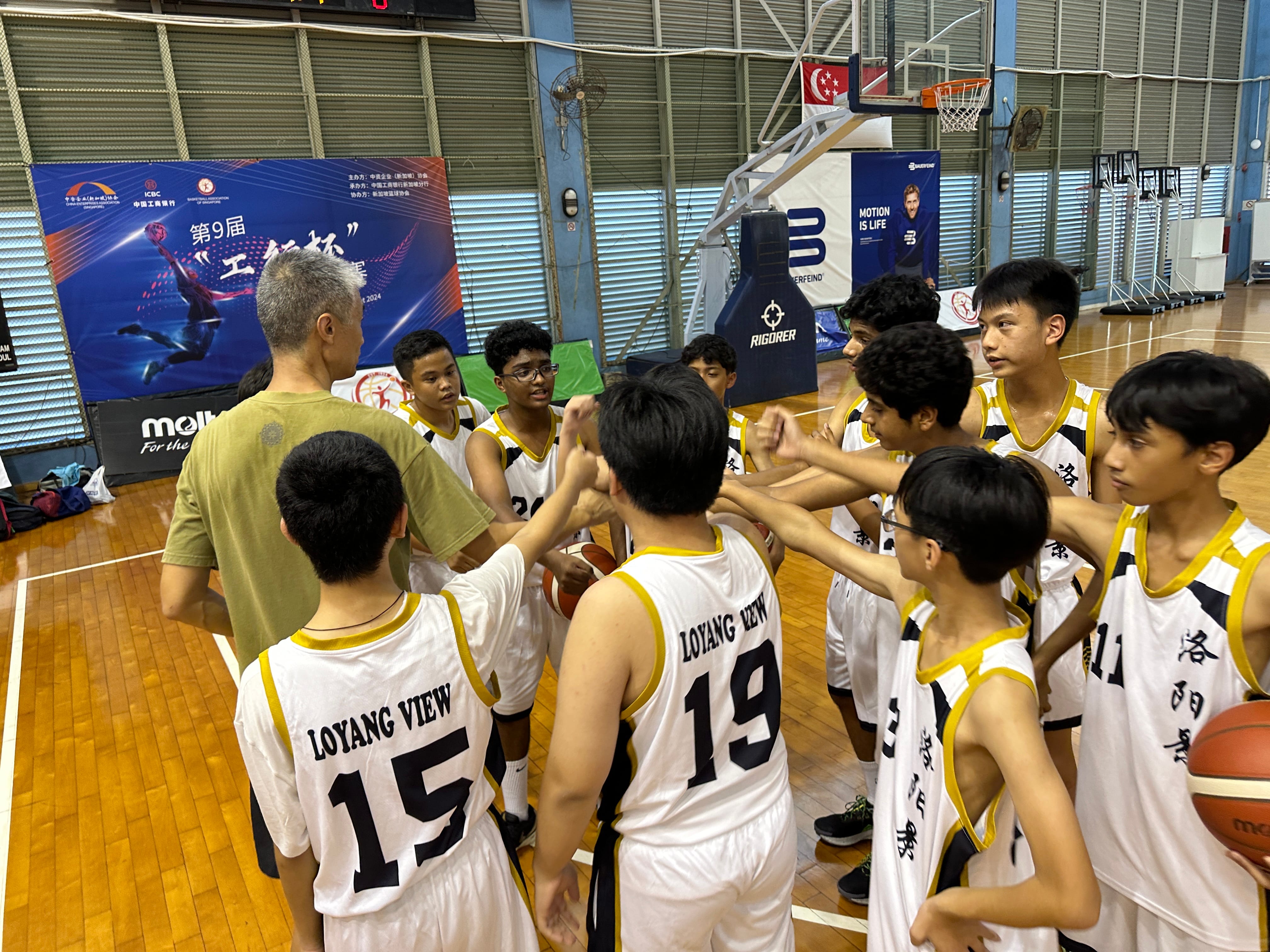 Youth basketball players in Loyang View jerseys huddle with a coach on an indoor court during a tournament.