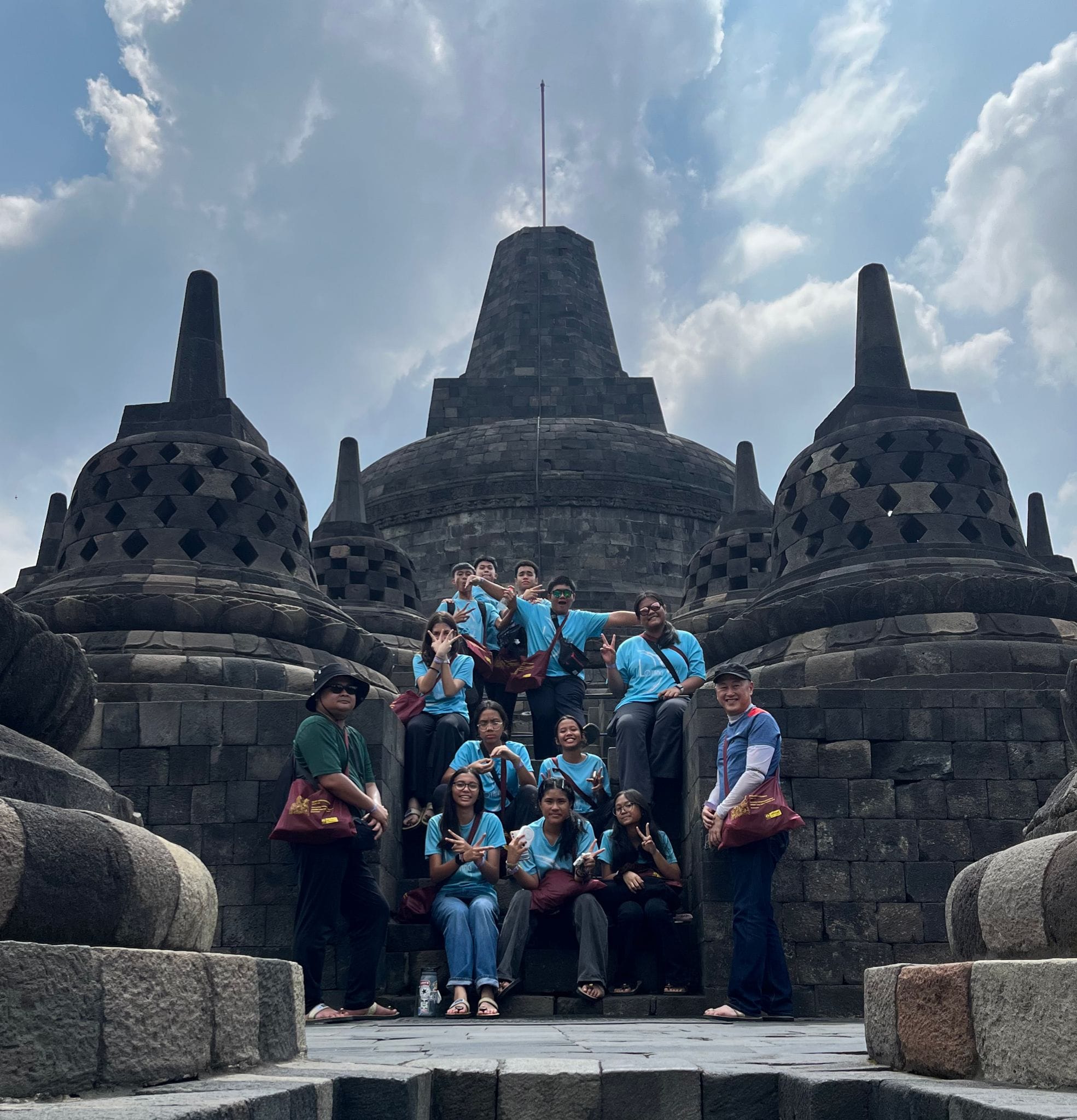 A group of young people in blue shirts pose on the stone steps of Borobudur temple with stupas behind them.