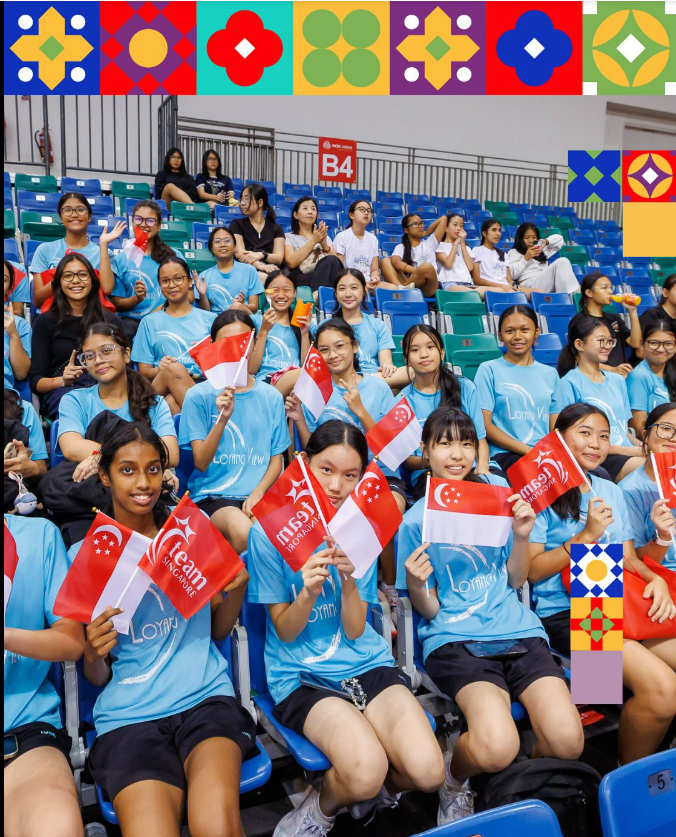 A large group of girls in light blue shirts wave Singapore flags while seated in stadium bleachers.