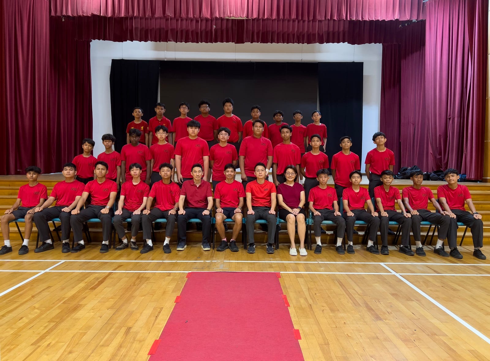 A large group of male students and teachers in red shirts pose in rows on a school hall stage.