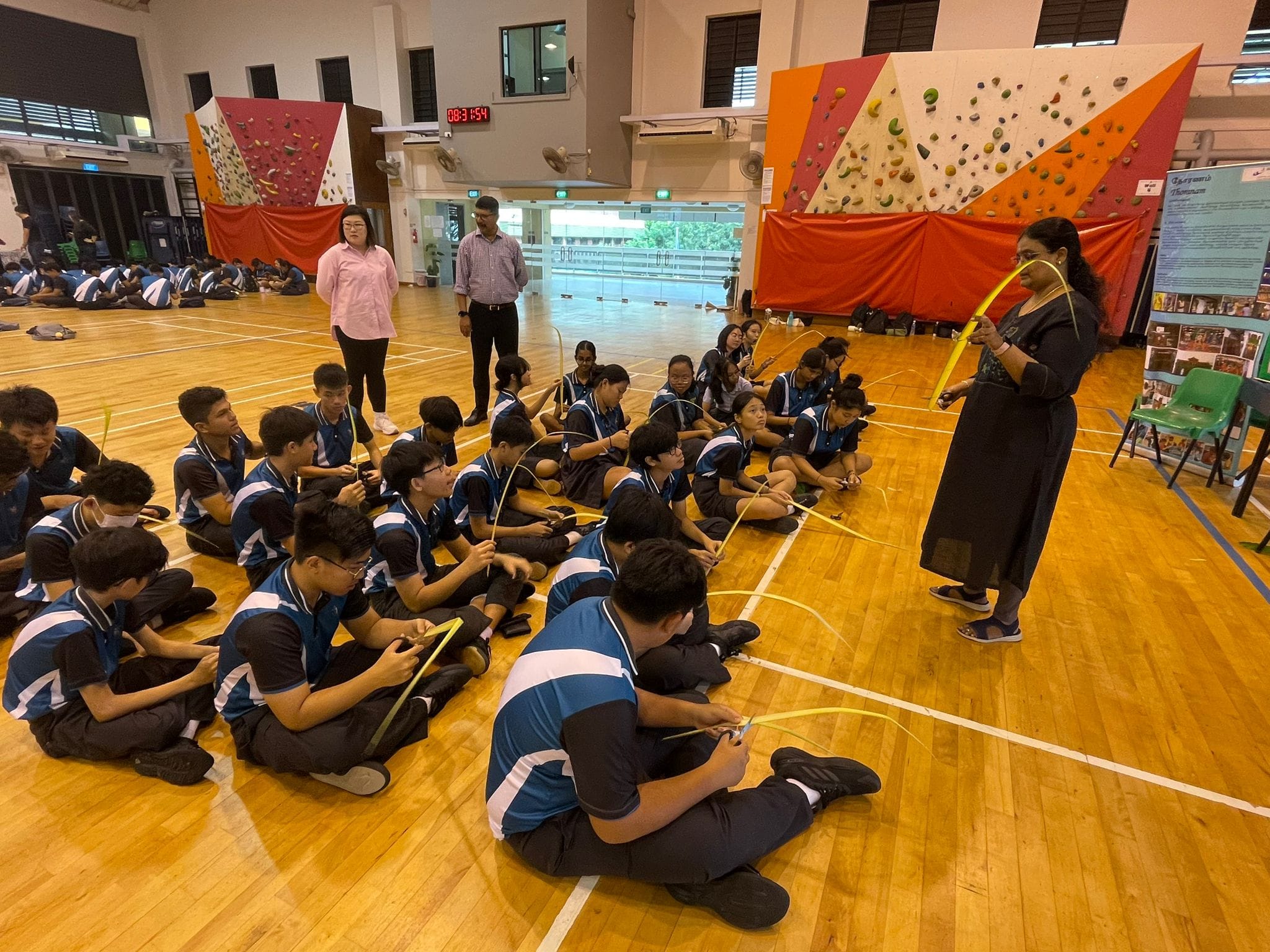 Students in blue uniforms sit on a gym floor holding yellow curved sticks while an instructor demonstrates in front of them.