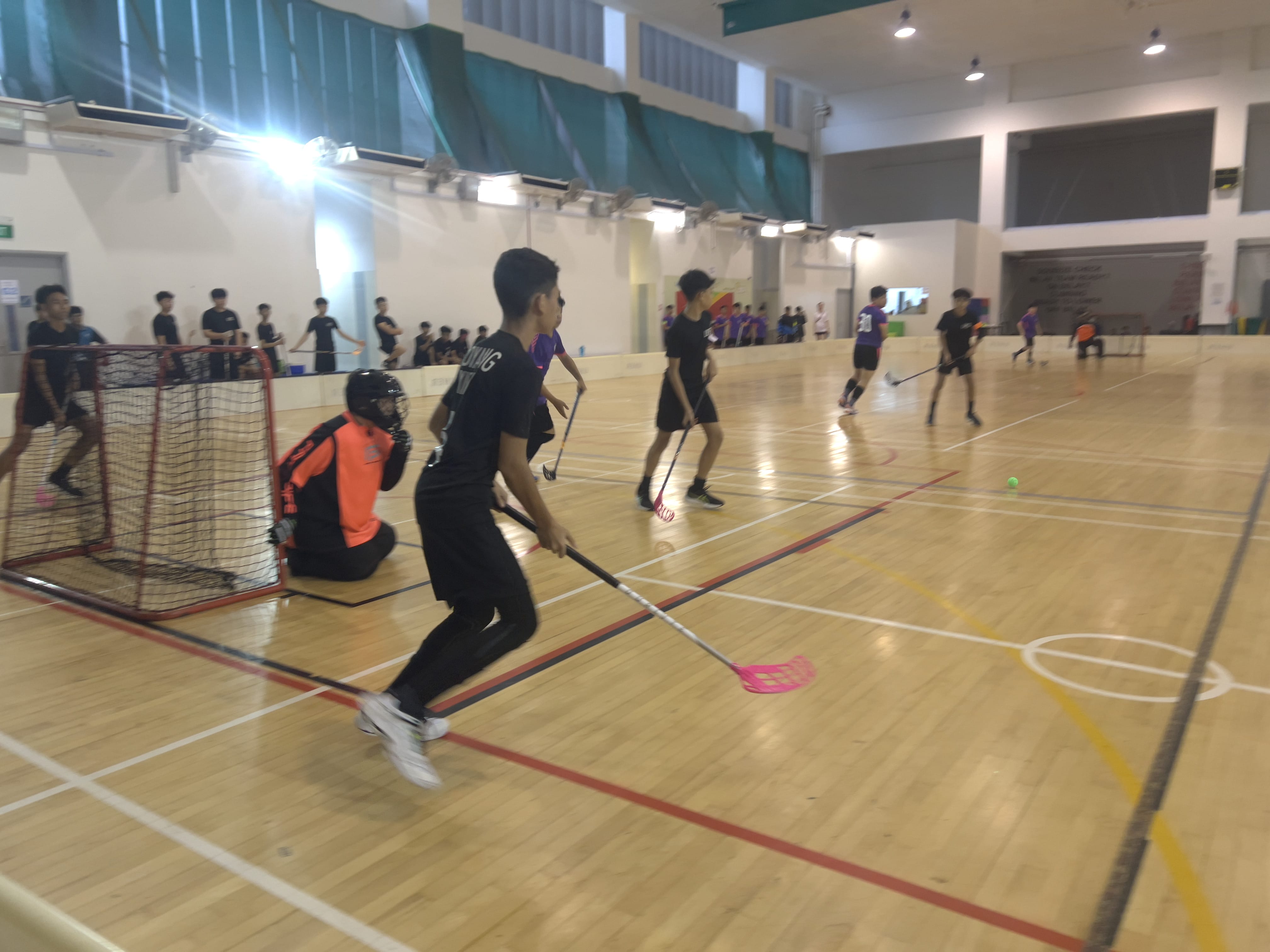 Players in black and purple compete in an indoor floorball match, with a goalie in orange guarding a red net.