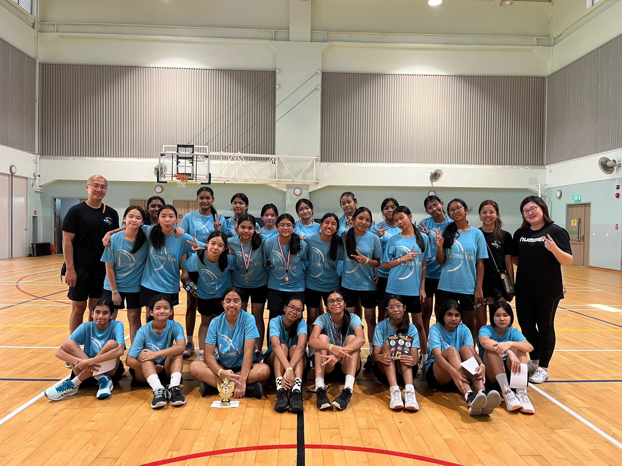A group of girls in light blue team shirts pose with a trophy and medals in an indoor basketball court.