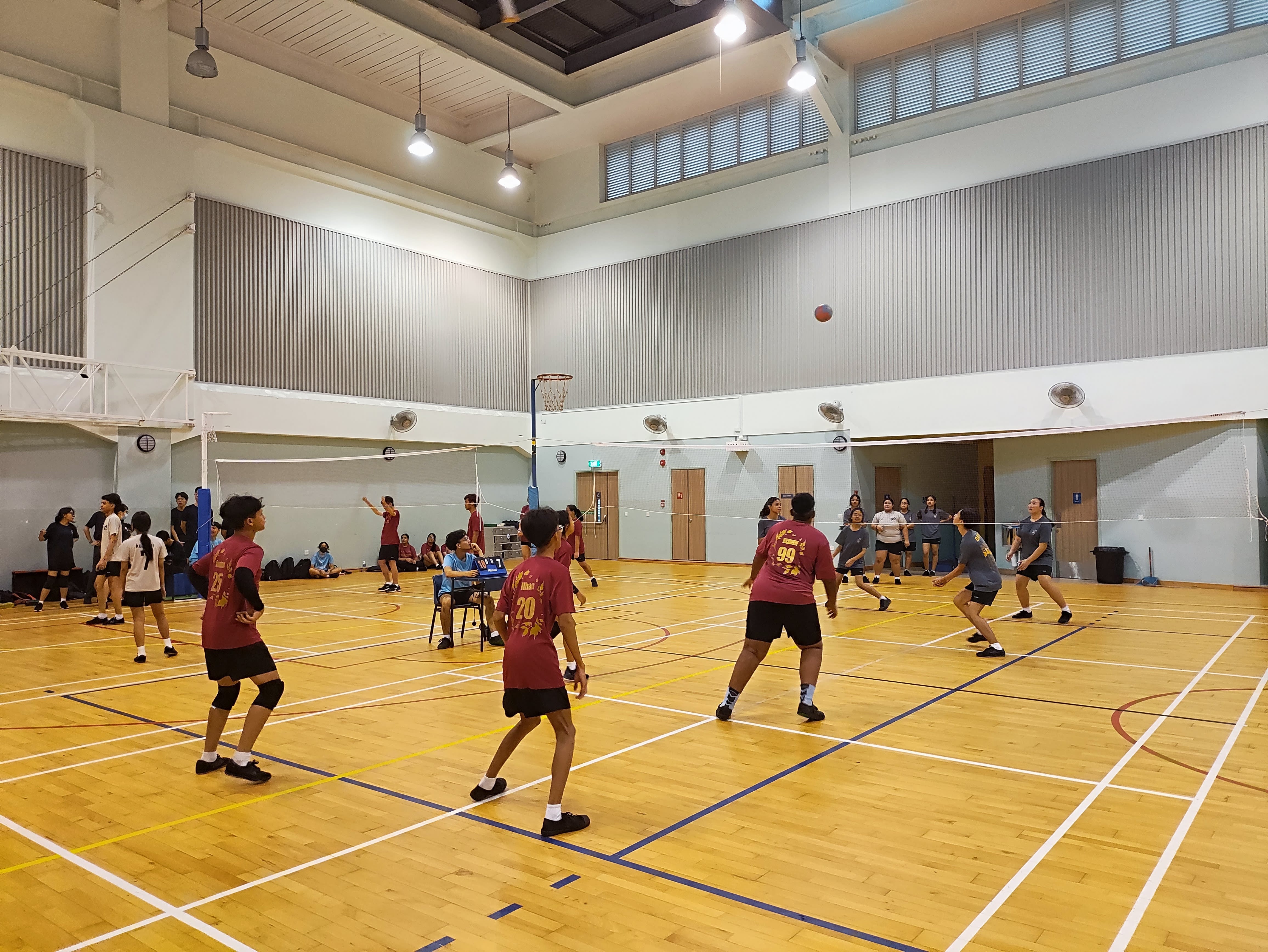Students in maroon jerseys play volleyball indoors on a wooden court with a net, ball mid-air, spectators seated nearby.