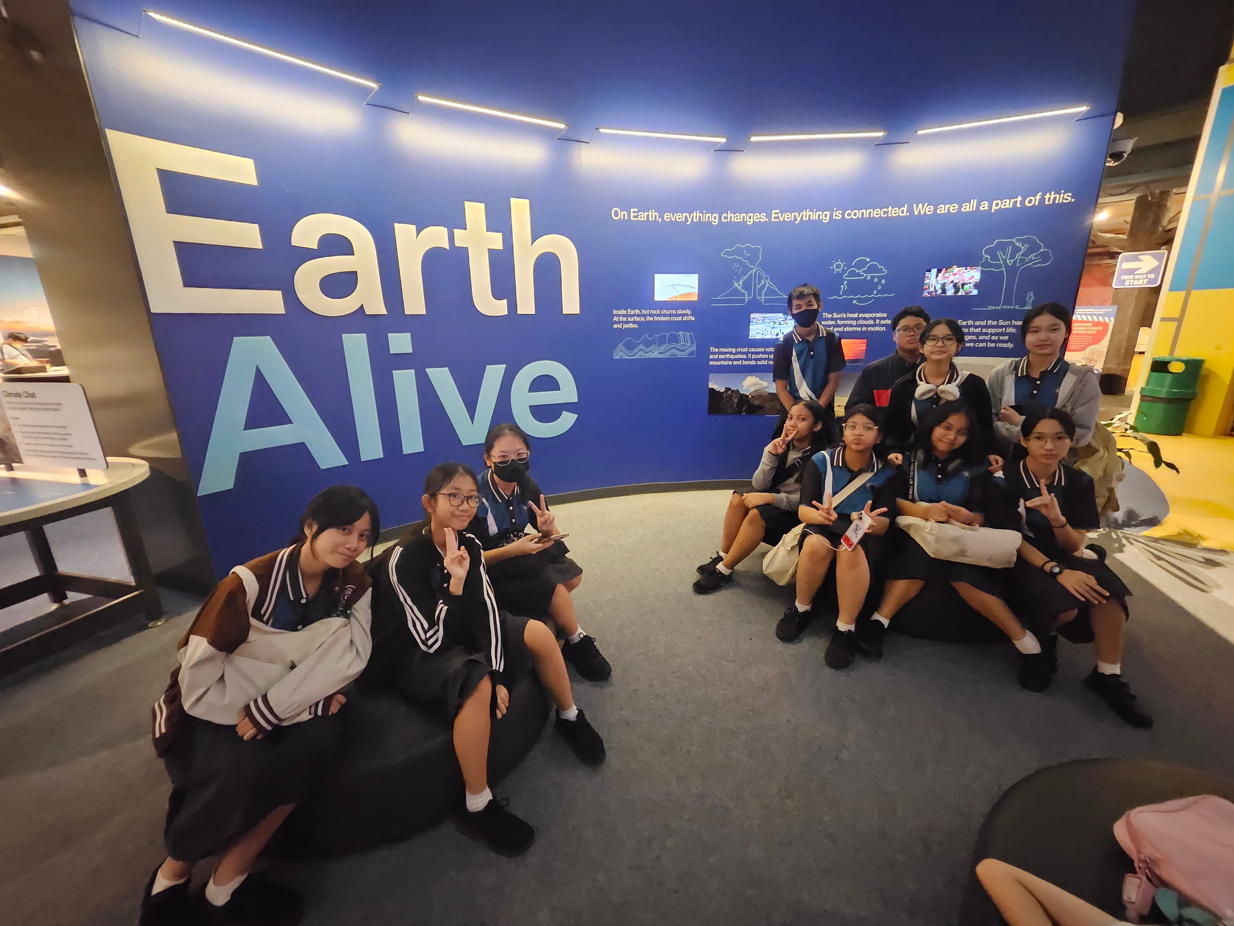 Students in school uniforms pose in front of a large blue Earth Alive museum exhibit display.