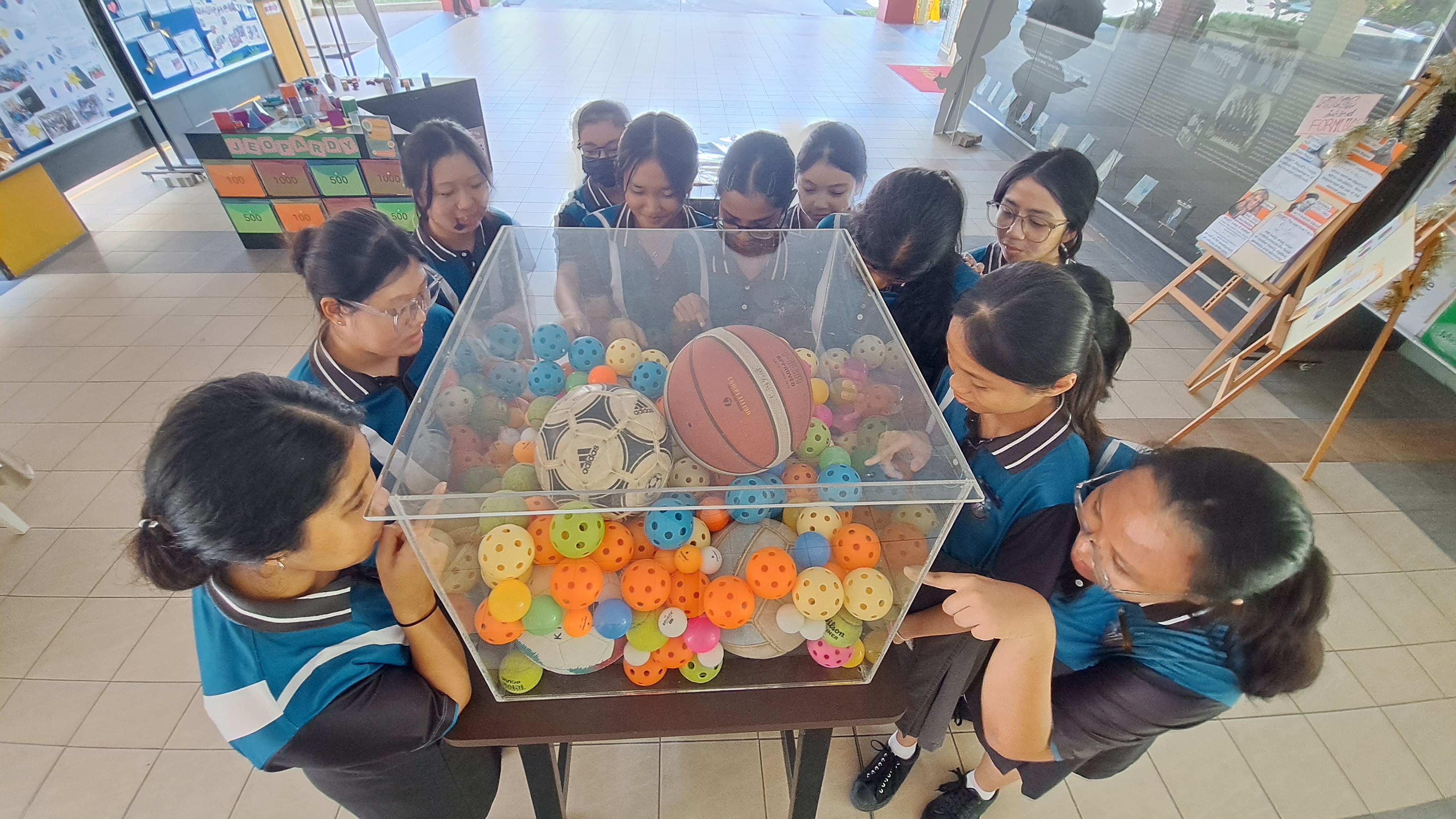 Students in blue uniforms gather around a clear box filled with colorful balls, a basketball, and a soccer ball.