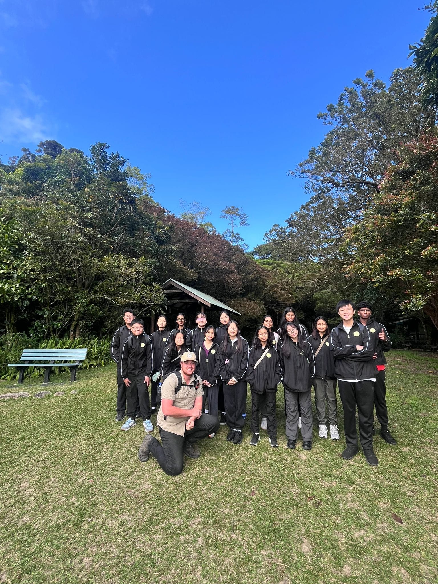 A group of young people in black tracksuits pose outdoors on grass with a guide kneeling in front, surrounded by trees.