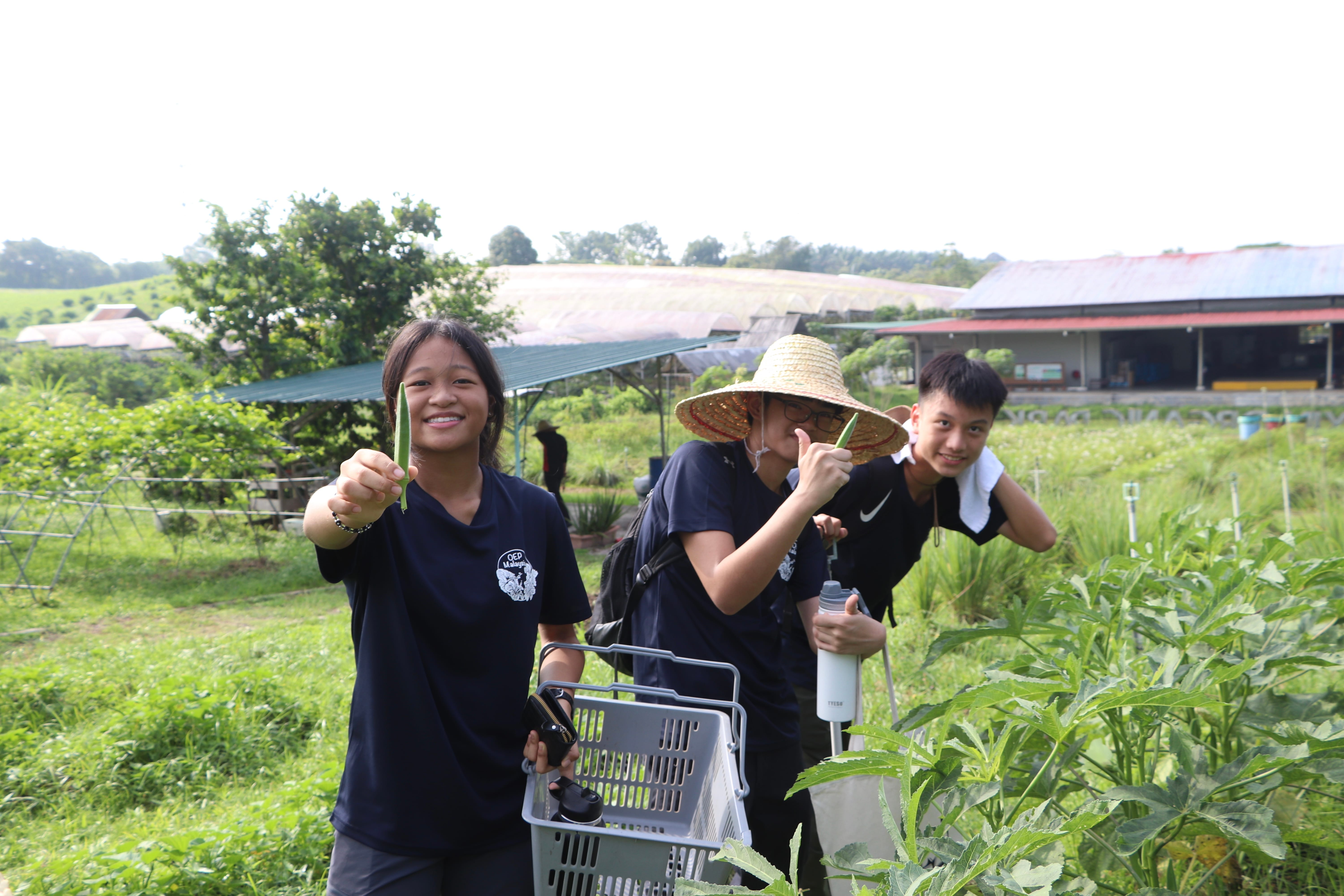 Three smiling young people stand among green farm plants, one holding a vegetable and another wearing a straw hat.