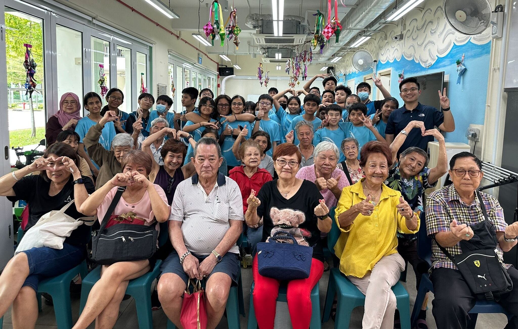 A large group of elderly adults and young volunteers in blue shirts pose cheerfully indoors with hand gestures.