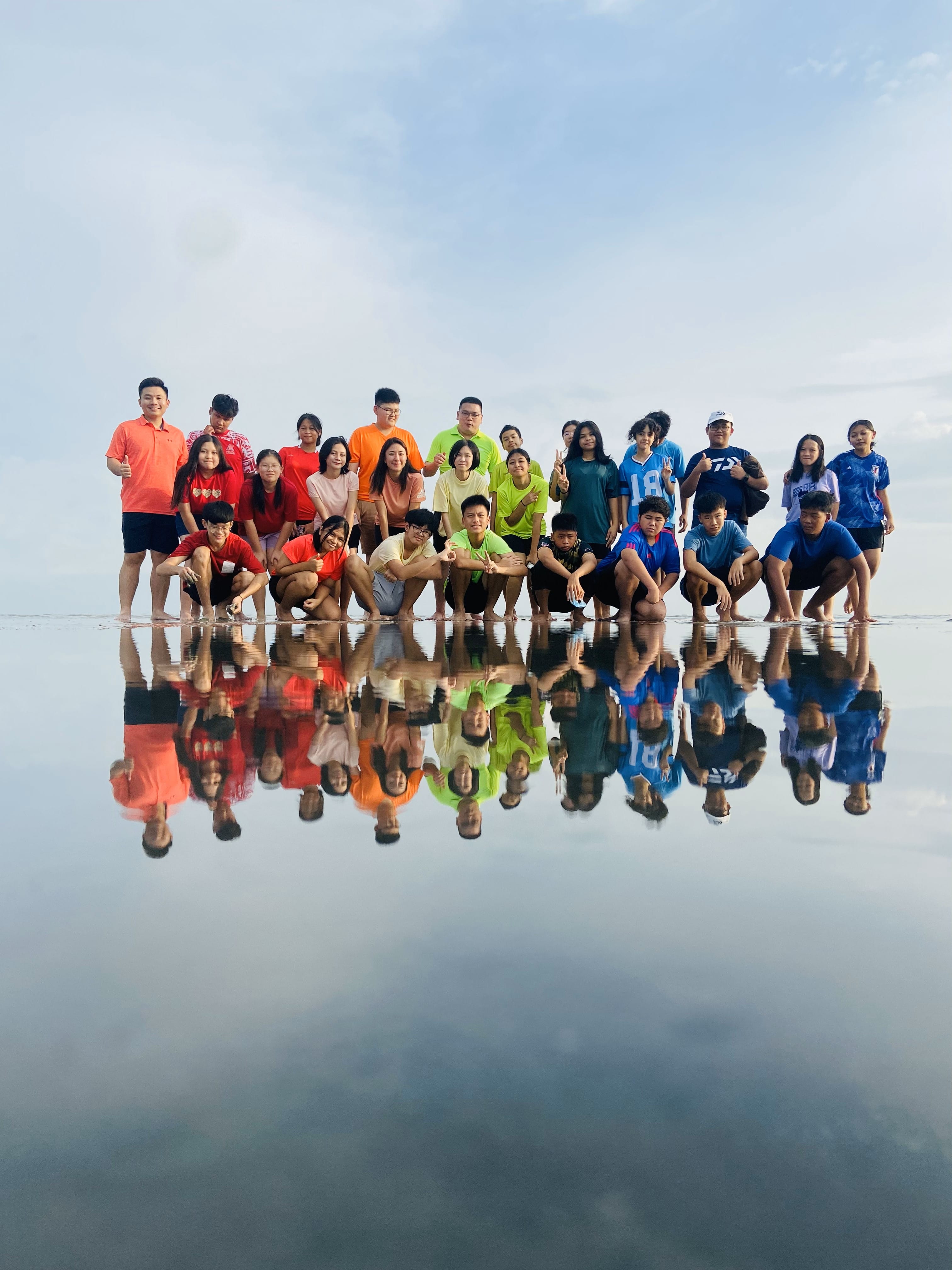 A large group of people posing on a reflective wet beach surface, their colorful shirts mirrored in the water below.