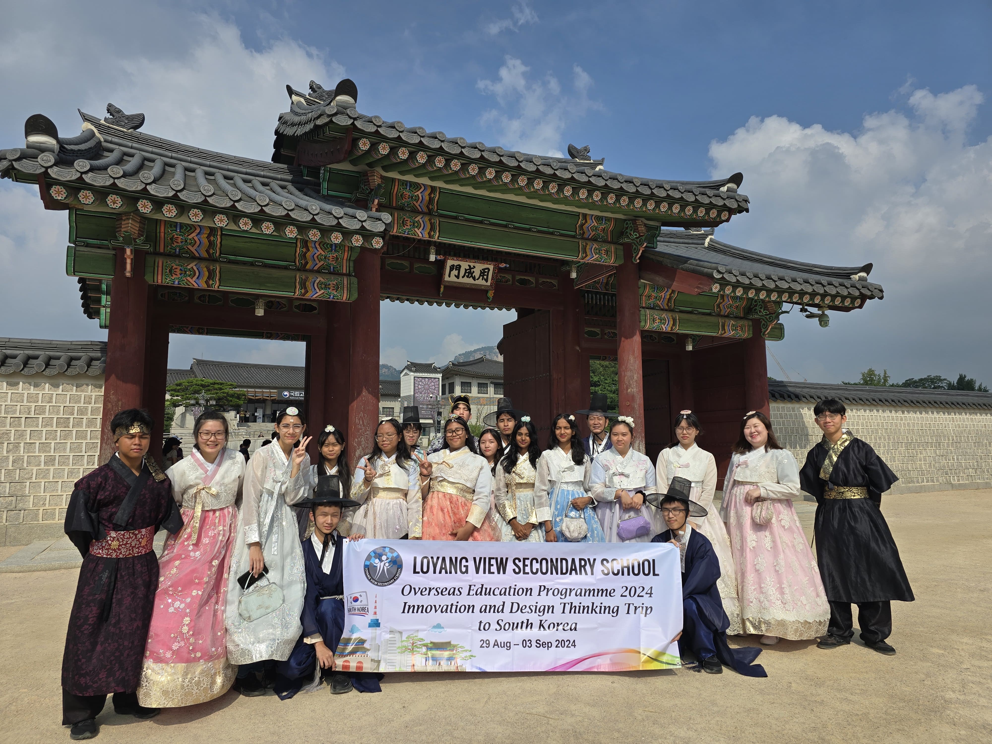 Group in hanbok at a Korean palace gate holding a Loyang View Secondary School South Korea trip 2024 banner.