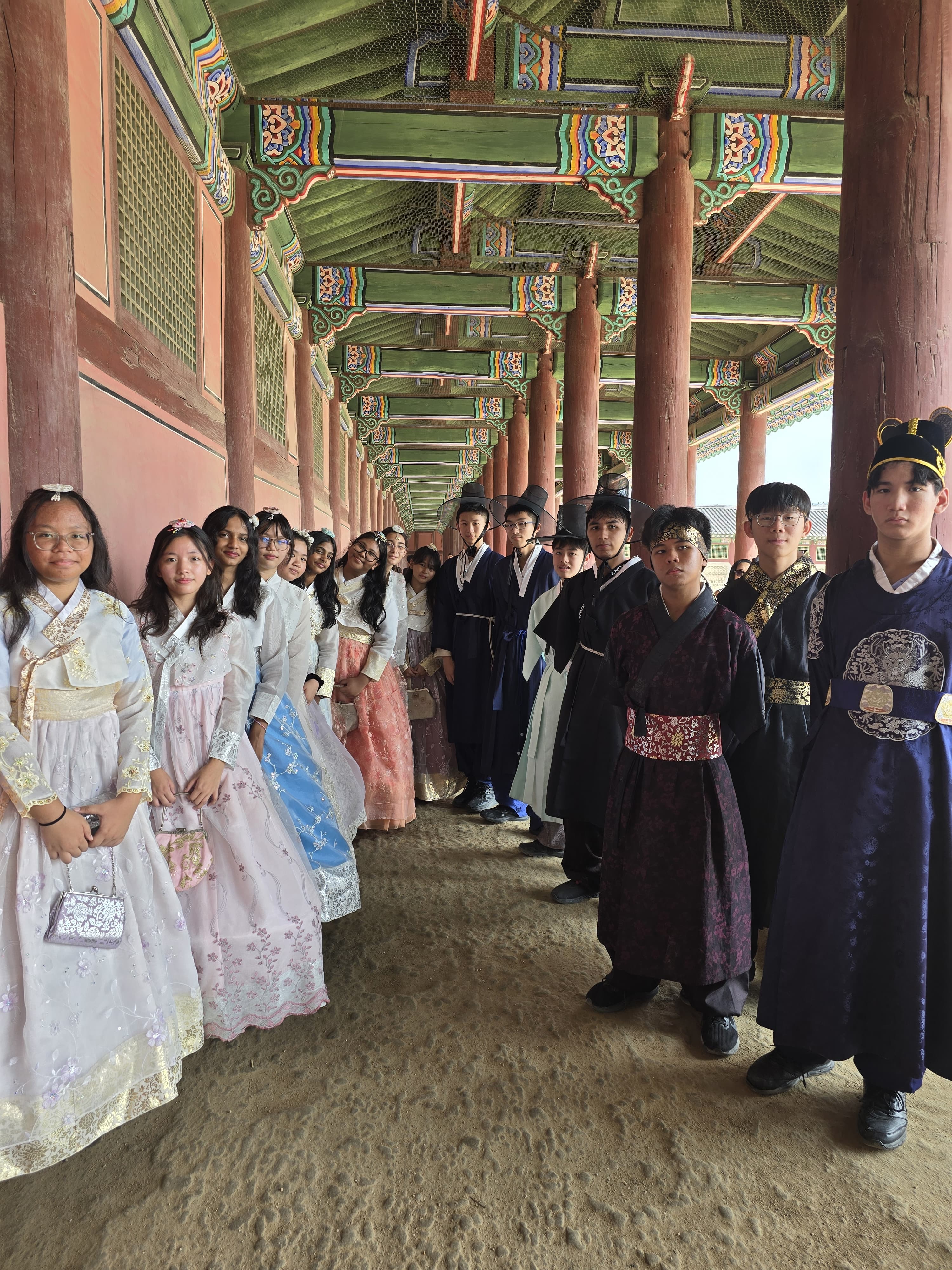 A group of young people wearing traditional Korean hanbok stand in a colorful palace corridor with red wooden columns.