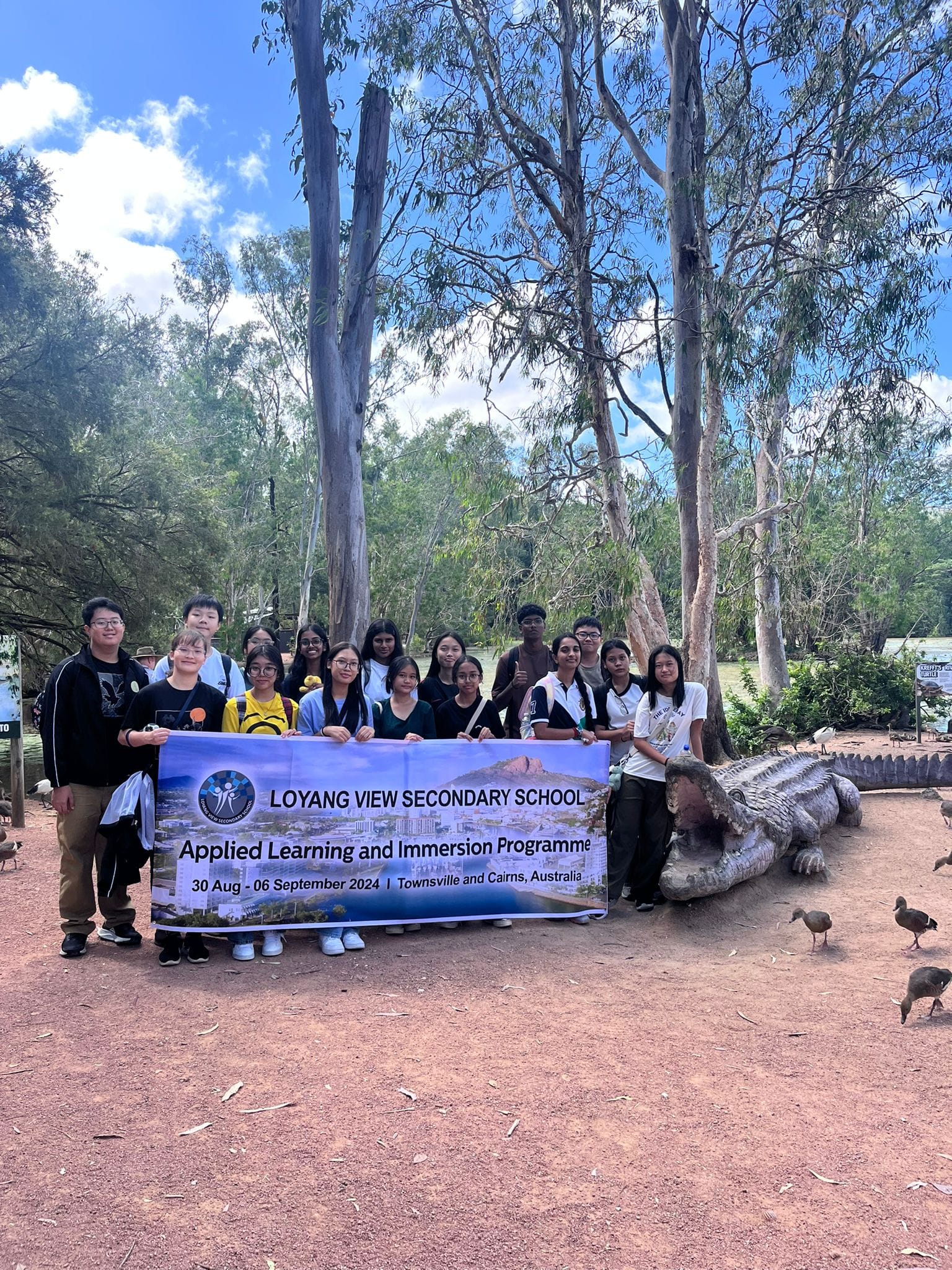 Students hold a Loyang View Secondary School banner at an outdoor Australian wildlife park with a crocodile statue nearby.