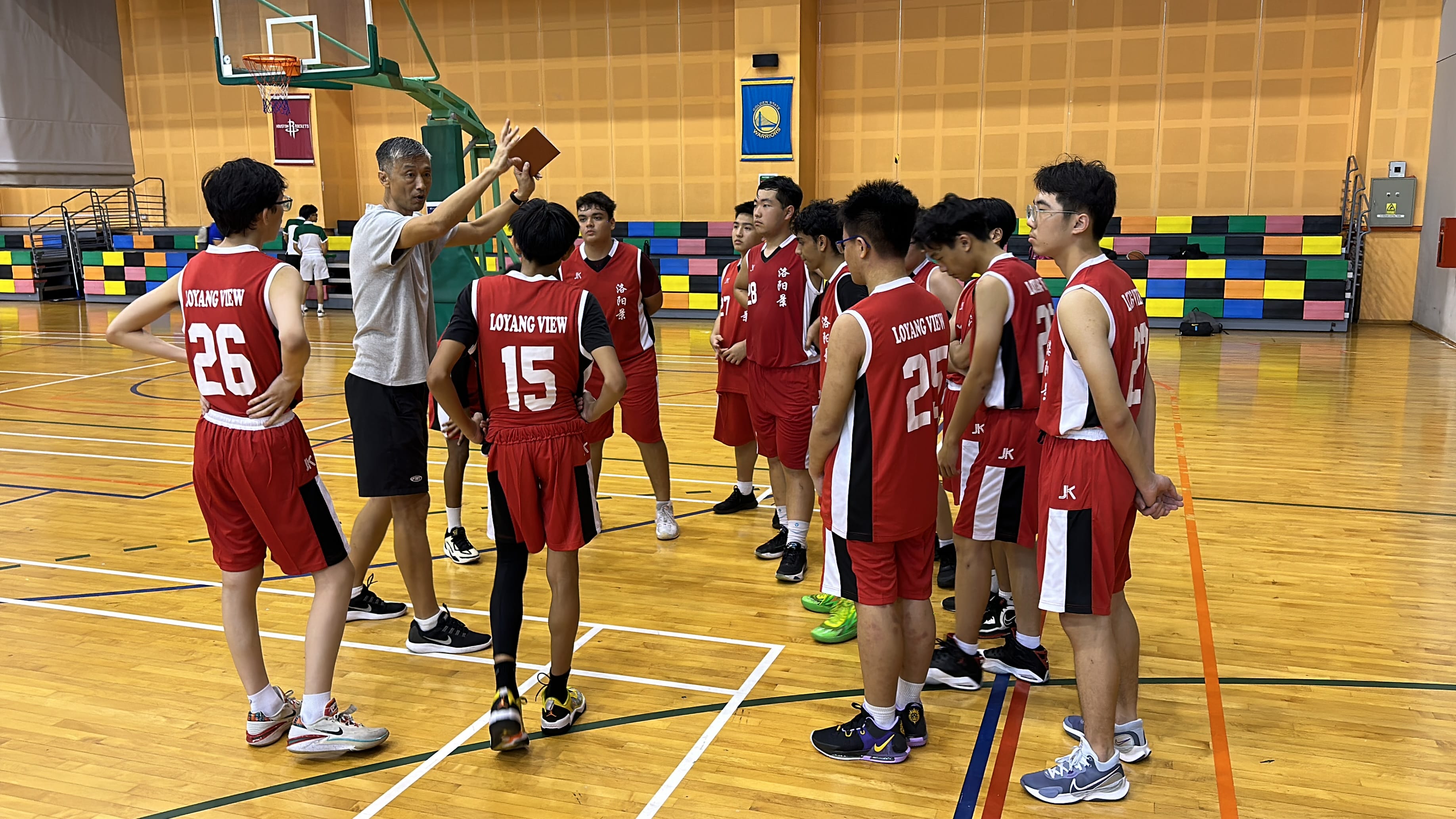 A coach gestures to a group of young players in red Loyang View jerseys during a basketball practice indoors.
