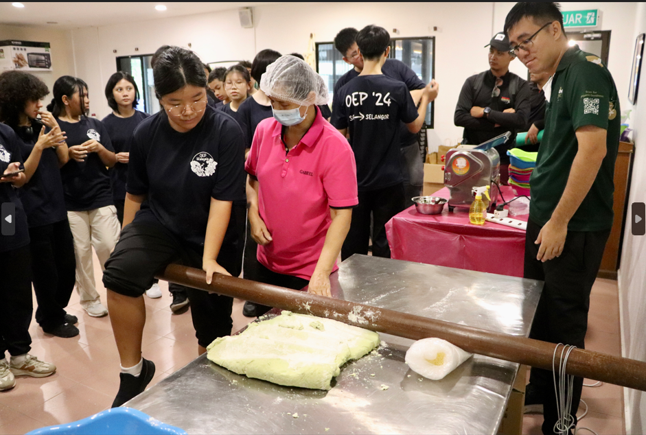 A student rolls green dough with a large rolling pin, guided by an instructor in a pink shirt, while classmates watch.