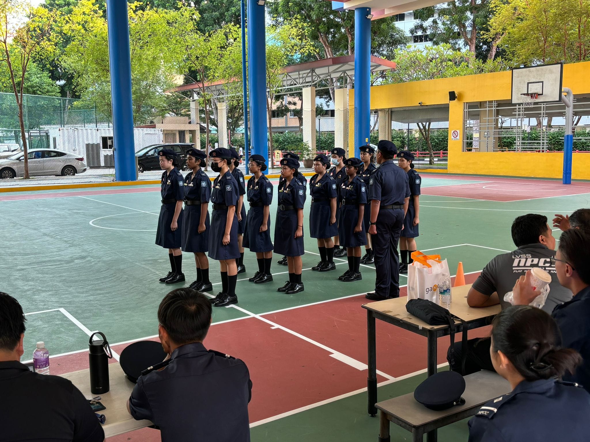 Students in navy blue uniforms and caps stand in formation on a colorful outdoor court while observers watch nearby.
