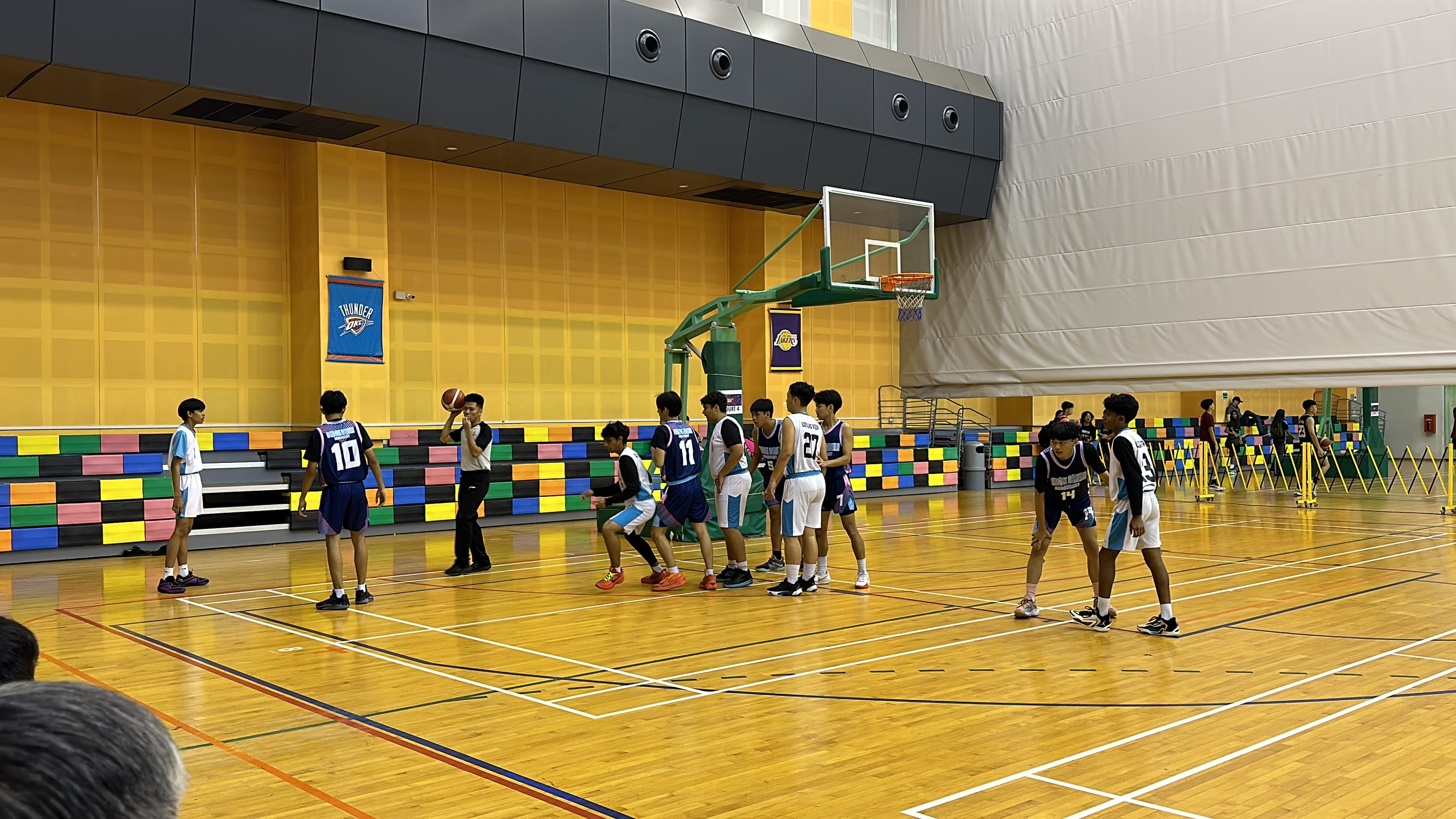 Young players in navy and white jerseys play basketball on an indoor hardwood court with a hoop and colorful bleachers.