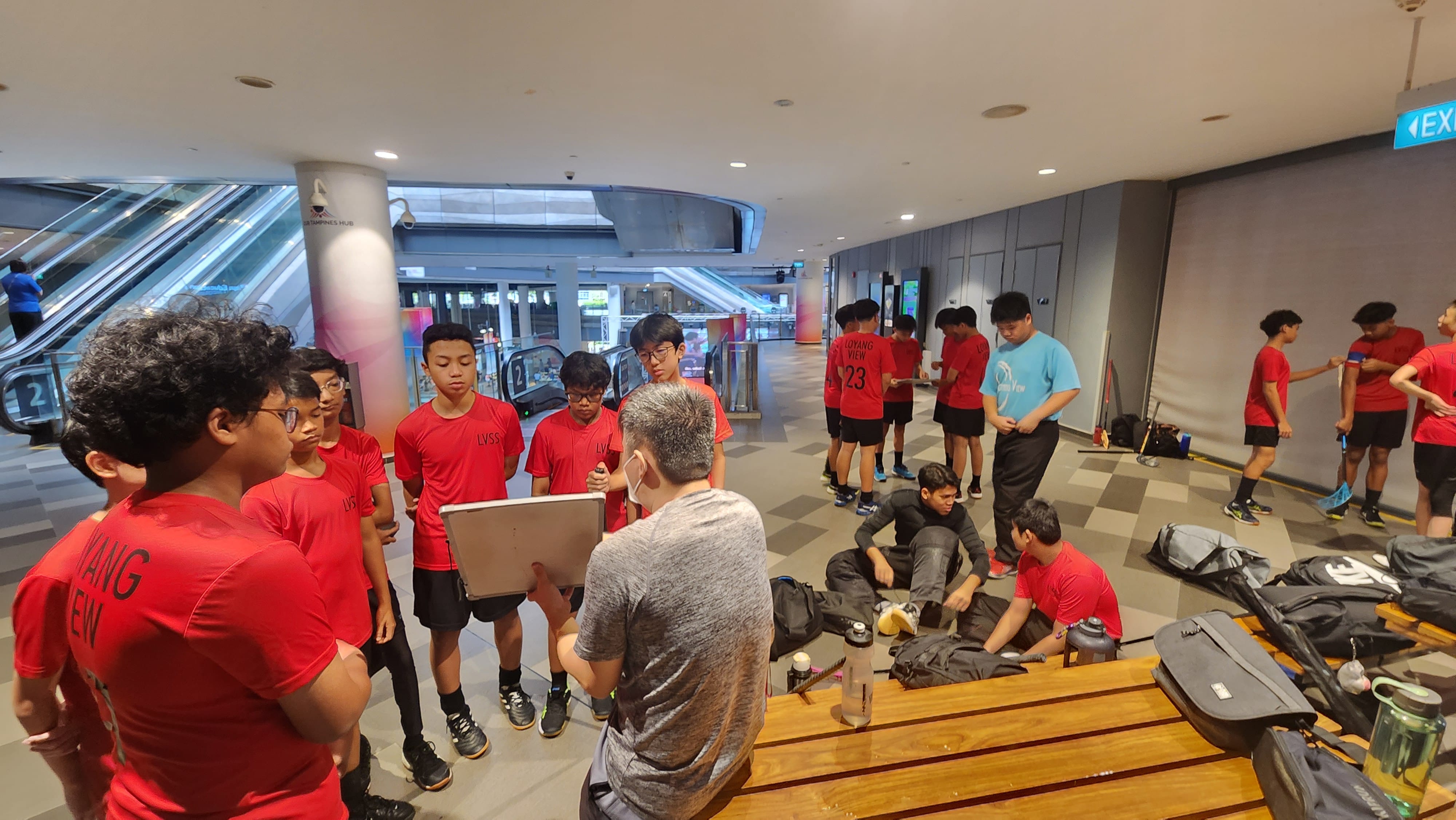 A coach shows a clipboard to teenage players in red jerseys gathered in an indoor building with escalators nearby.