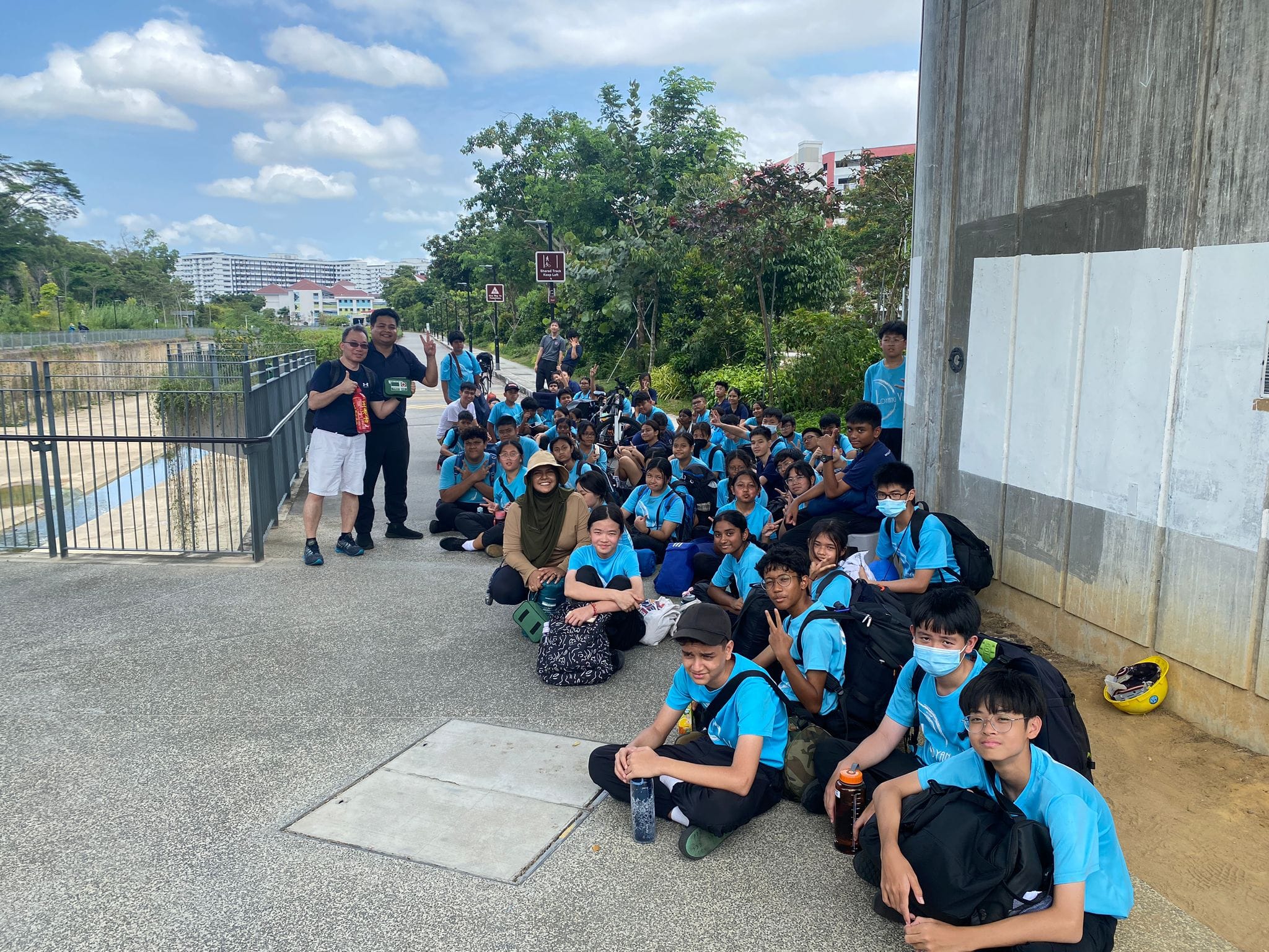 A large group of students in blue shirts sit and stand on a pathway outdoors near a wall and trees.