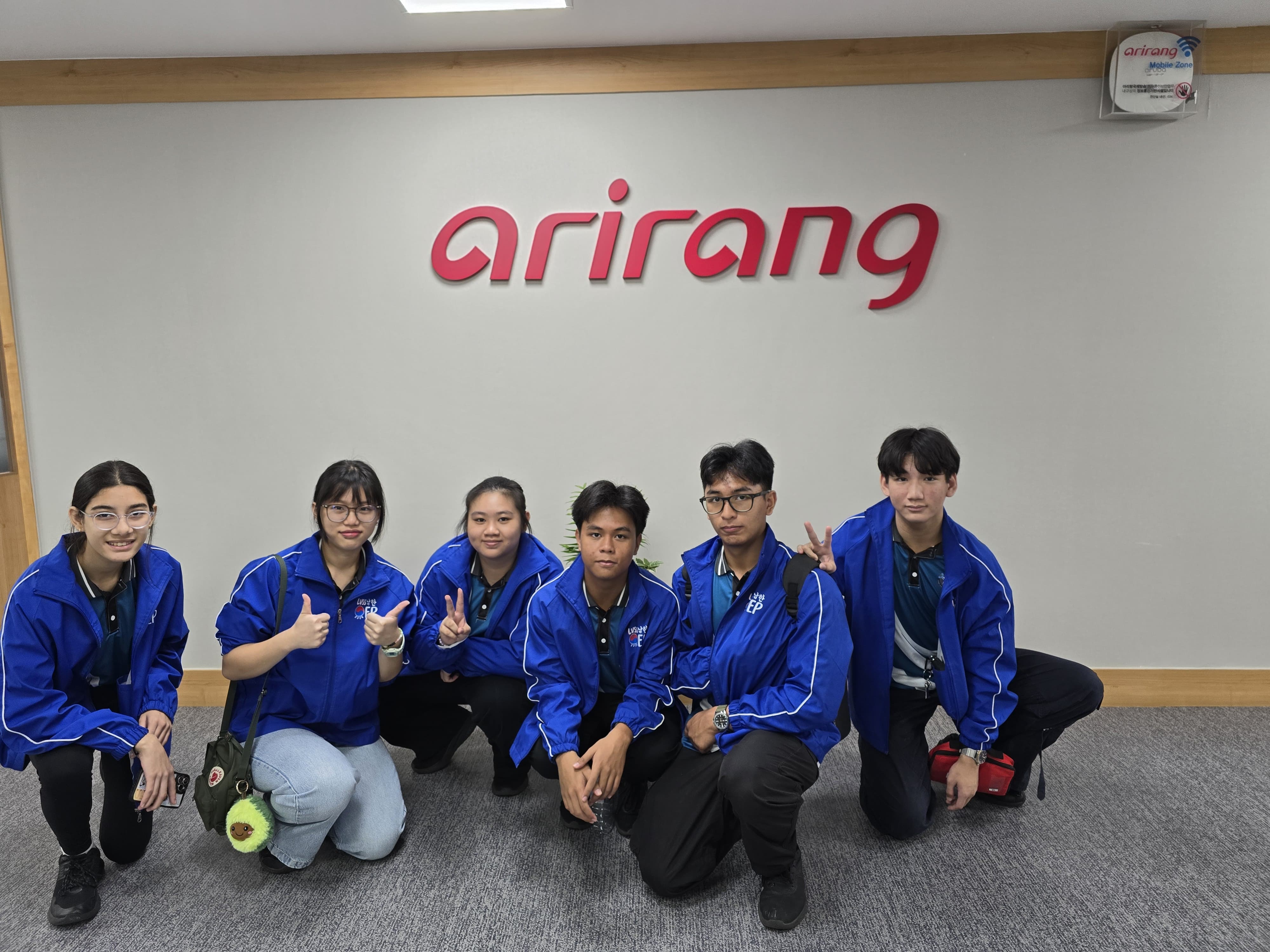 Six young people in matching blue jackets pose crouching in front of a wall with the red Arirang sign.