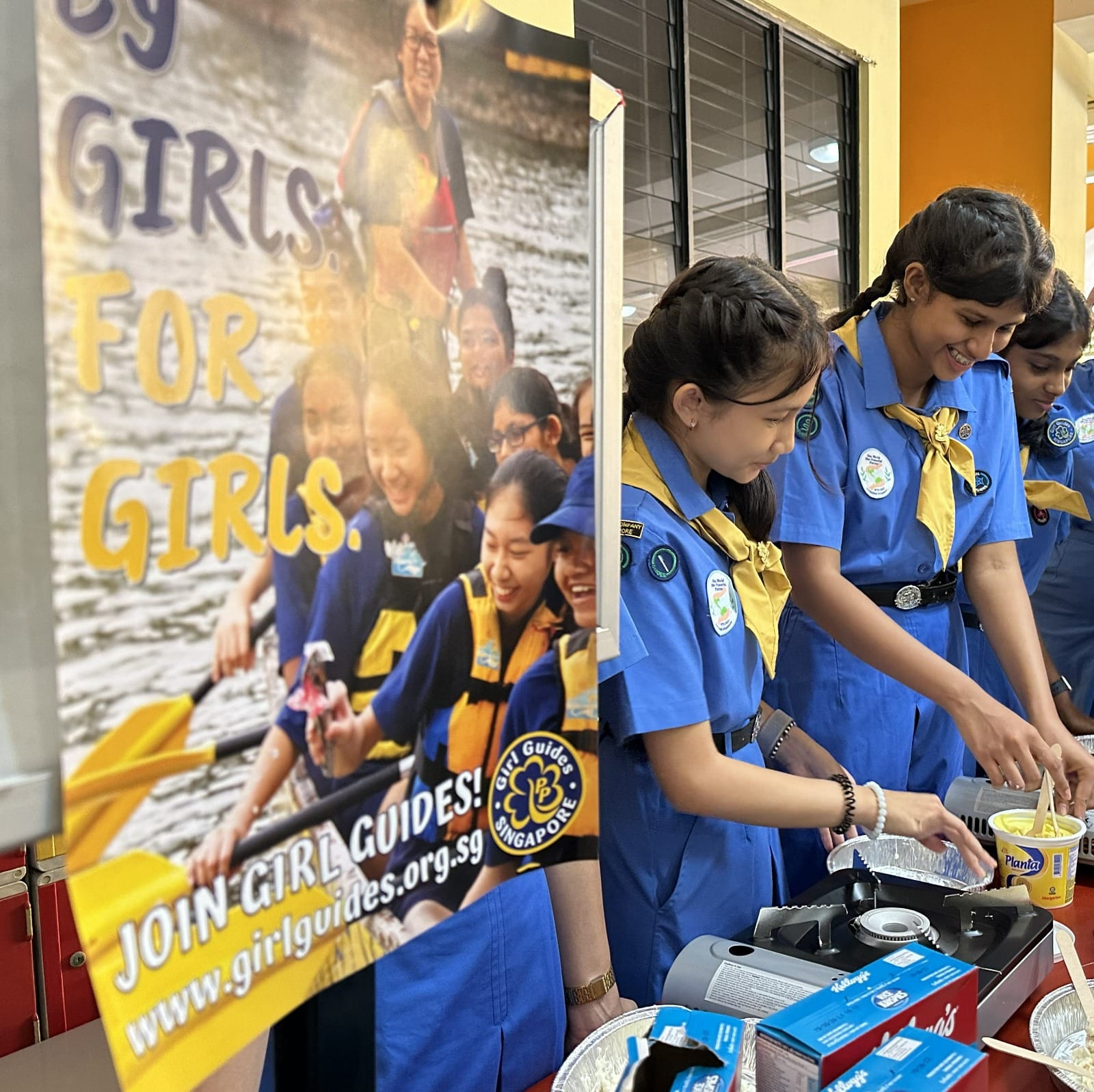 Girl Guides Singapore members in blue uniforms cooking at a table beside a recruitment banner.