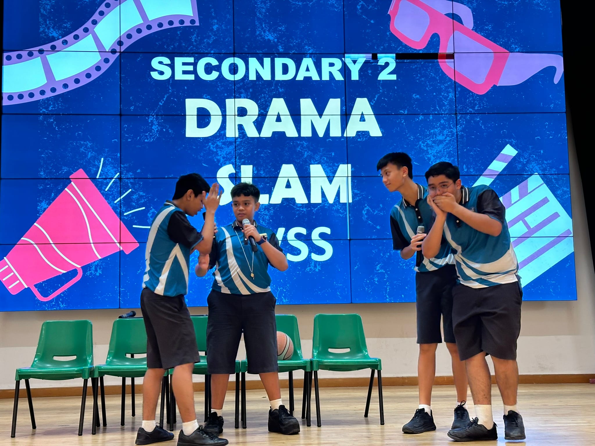 Four students in blue uniforms perform on stage at a Secondary 2 Drama Slam event, two holding microphones.