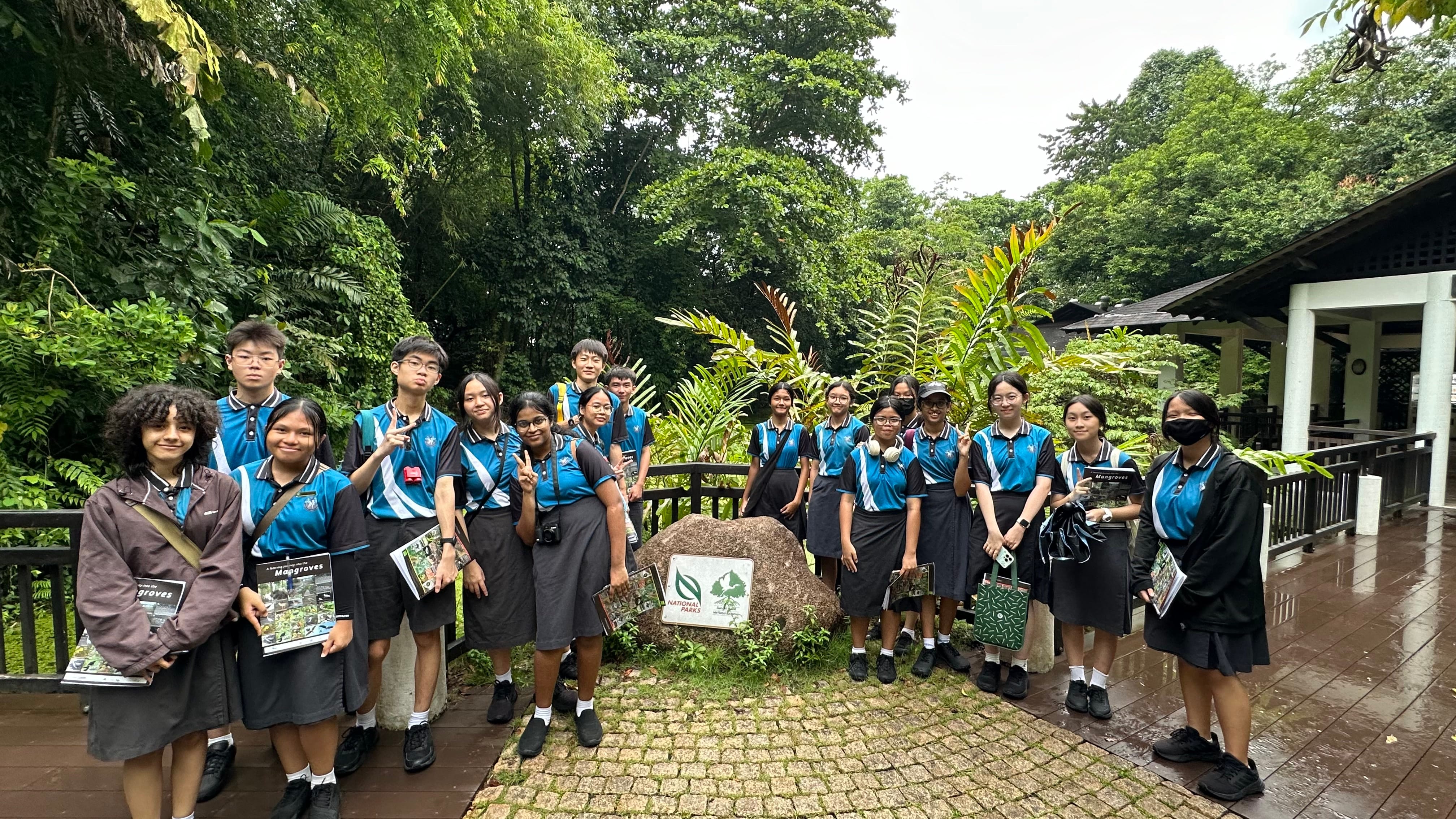 A group of students in blue uniforms pose near a National Parks boulder sign surrounded by lush green trees.