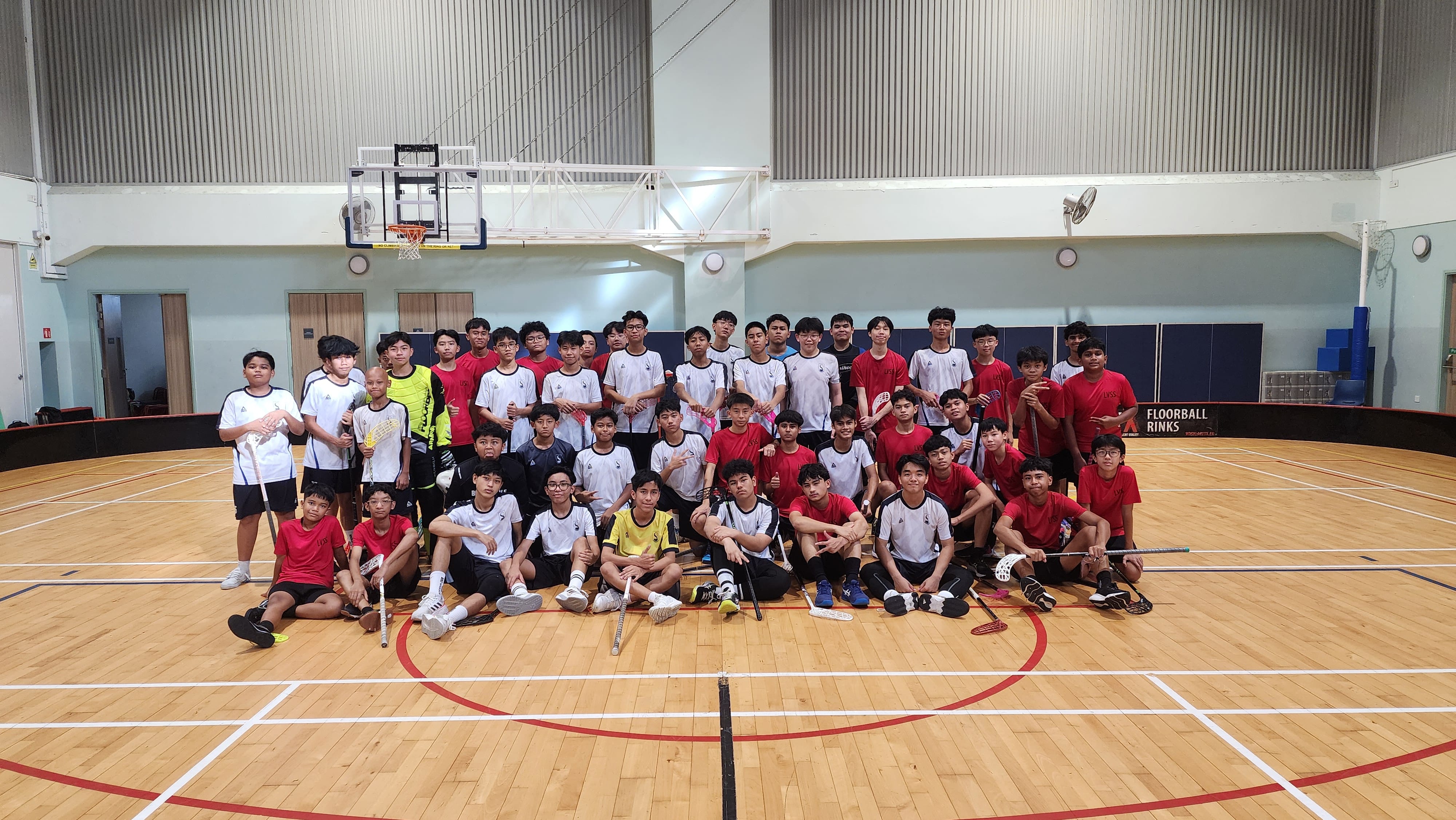 A large group of boys in red and white jerseys pose with floorball sticks on an indoor sports court.