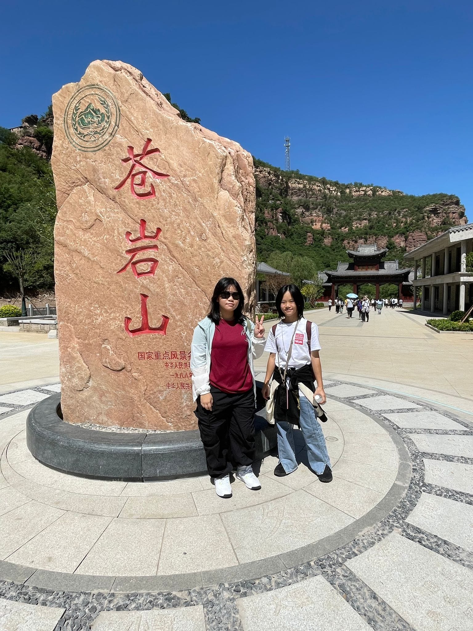 Two young women pose beside a large stone monument with red Chinese characters at a scenic mountain park.