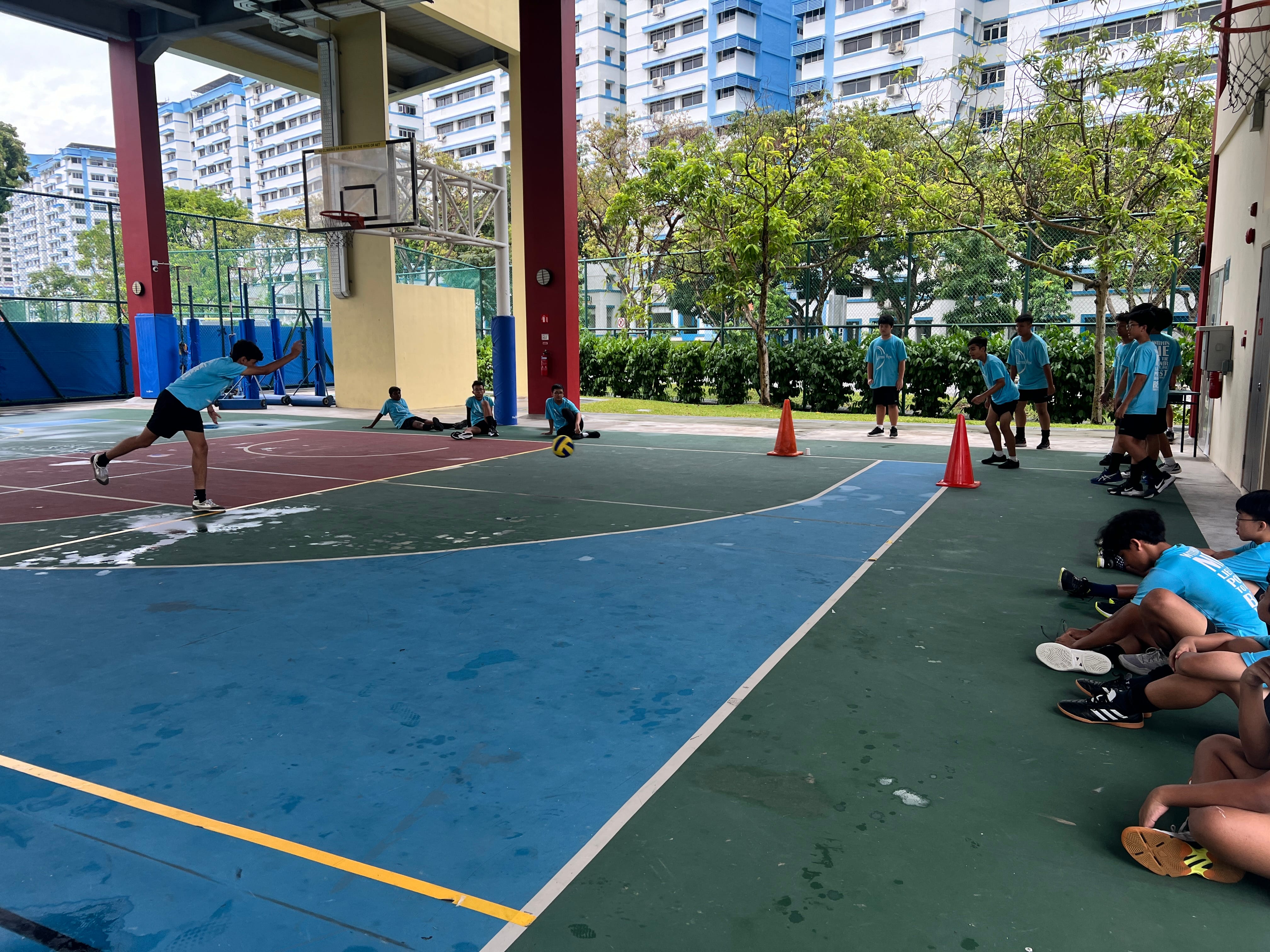 Boys in blue shirts play on an outdoor court as one throws a ball; others watch near orange cones.