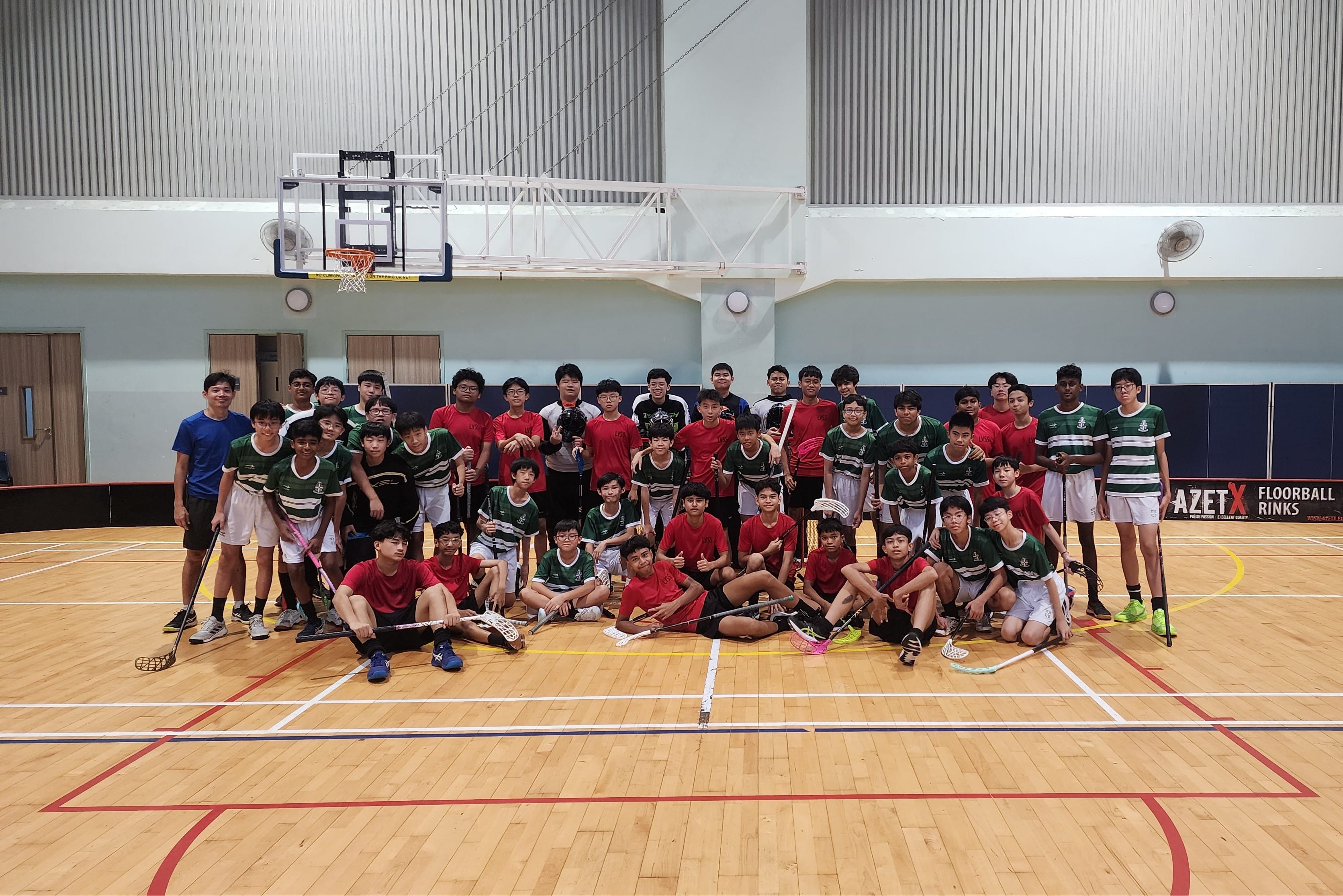 A large group of boys in red and green jerseys pose with floorball sticks inside an indoor sports hall.