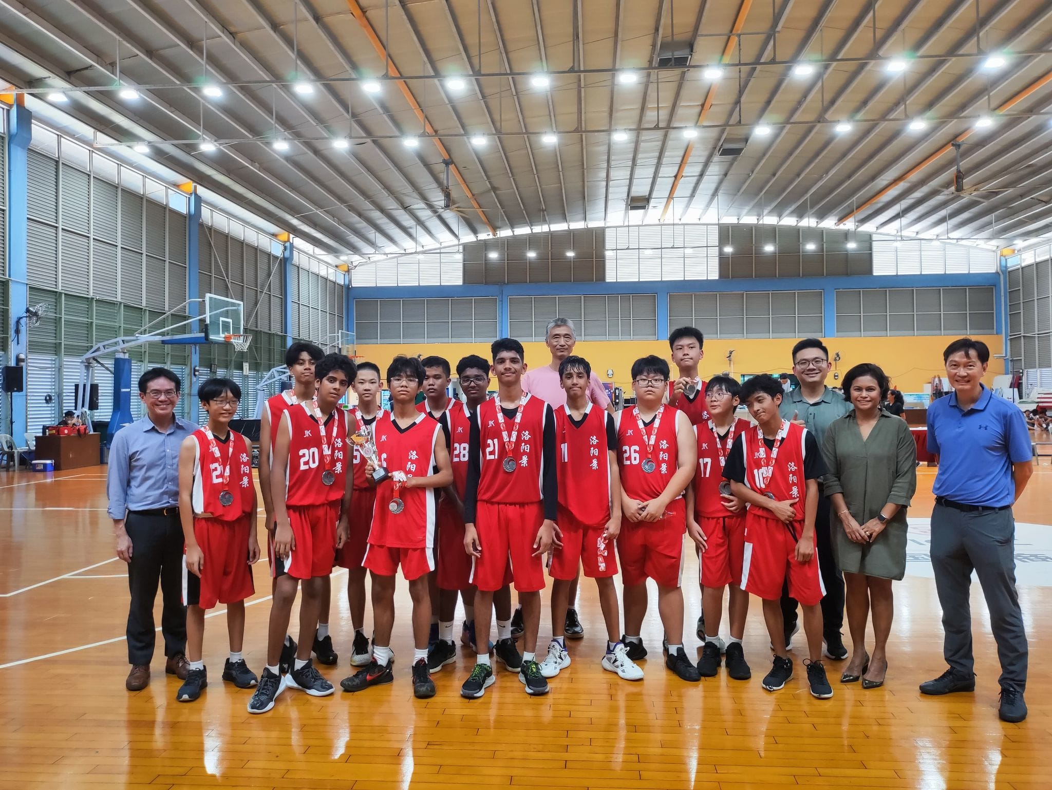 Youth basketball team in red uniforms with medals poses with adults on an indoor court after a tournament.