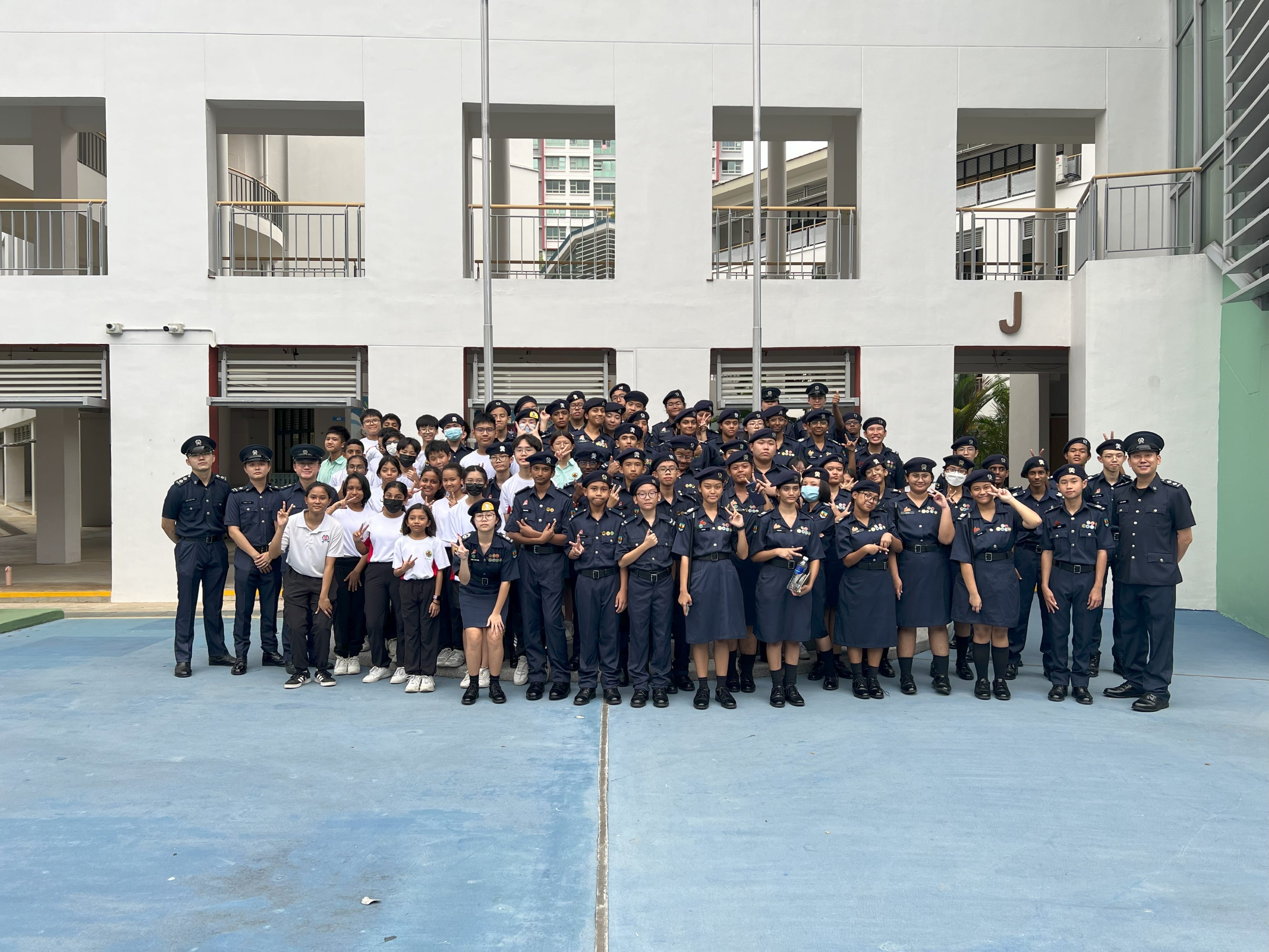 Large group of students mostly in dark navy uniforms posing outdoors in front of a white multi-storey school building.