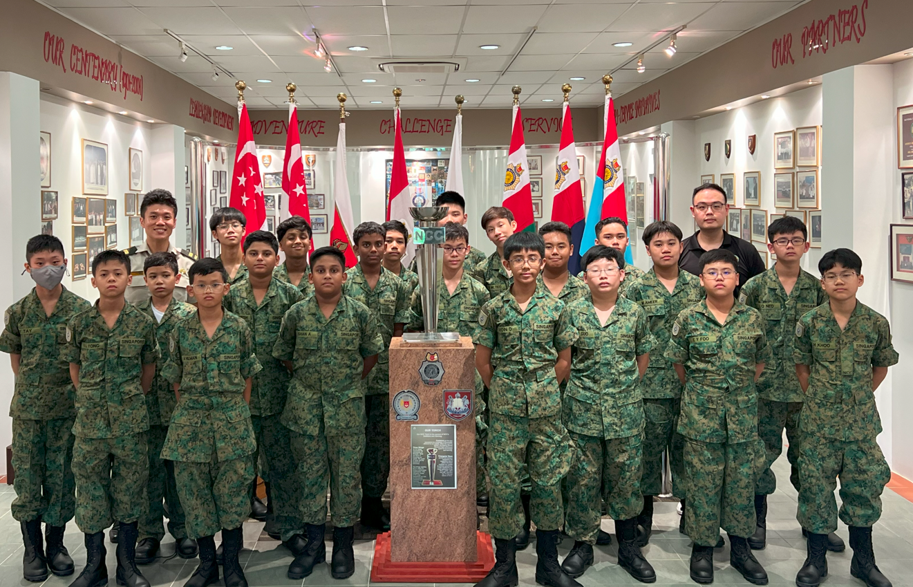 A group of young cadets in camouflage uniforms pose around a trophy pedestal with flags and plaques in a hall.