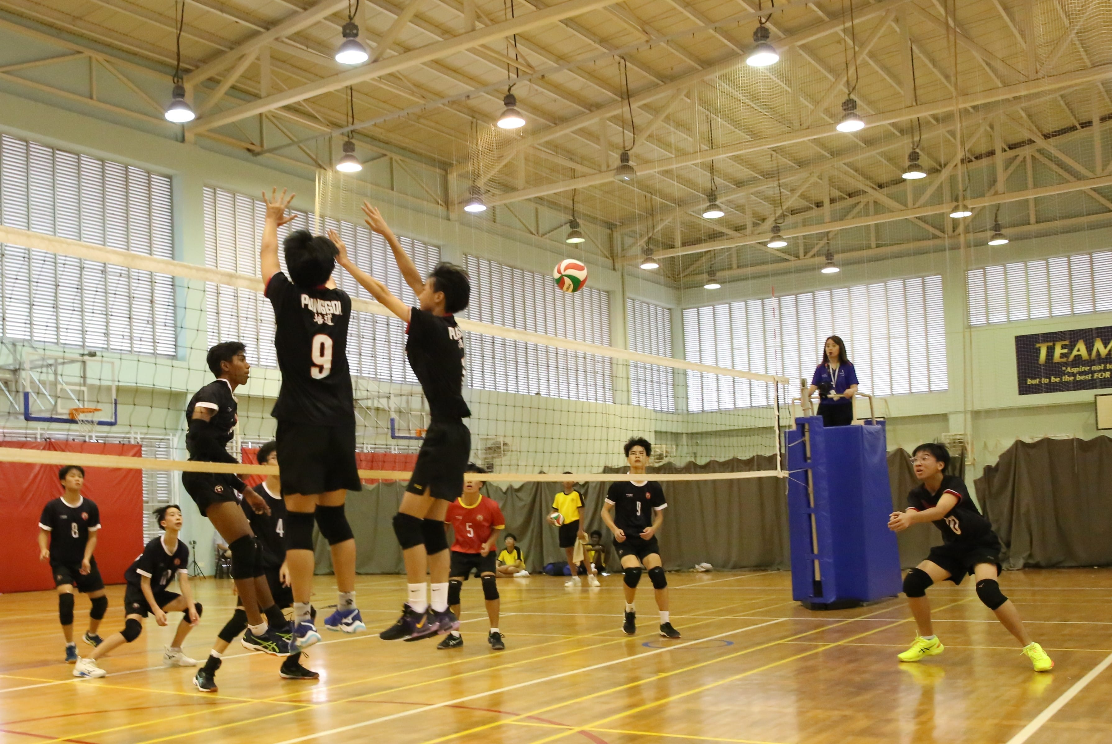 Two players in black jerseys jump to block at the net during an indoor youth volleyball match.